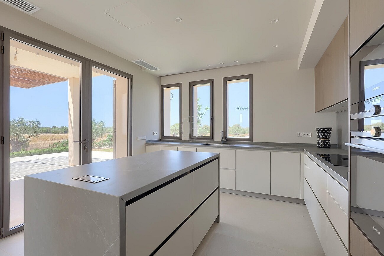 Modern kitchen with large central island, white cabinetry, integrated appliances, and view through glass doors to patio.