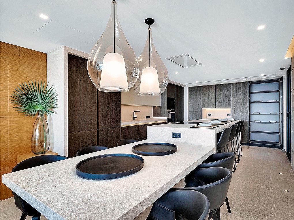 Modern open-plan kitchen featuring a long light-colored stone island, dark cabinetry, and two clear glass pendant lights.