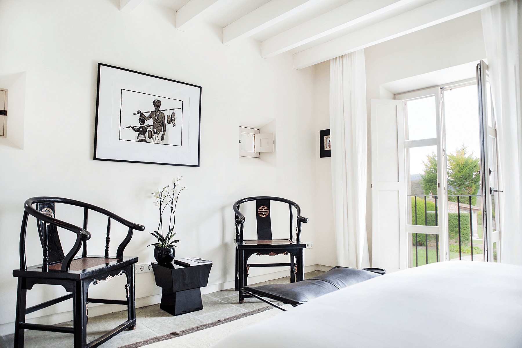 A bright living space with white walls, beamed ceilings, and two dark wooden antique Chinese-style chairs flanking a side table.