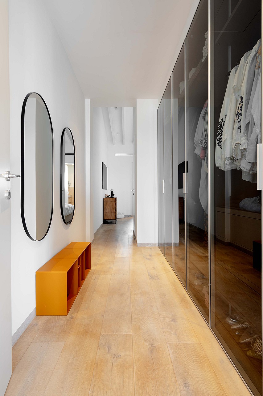 A bright hallway featuring light wood flooring, a long wall of dark glass-fronted wardrobes, and minimalist wall mirrors.