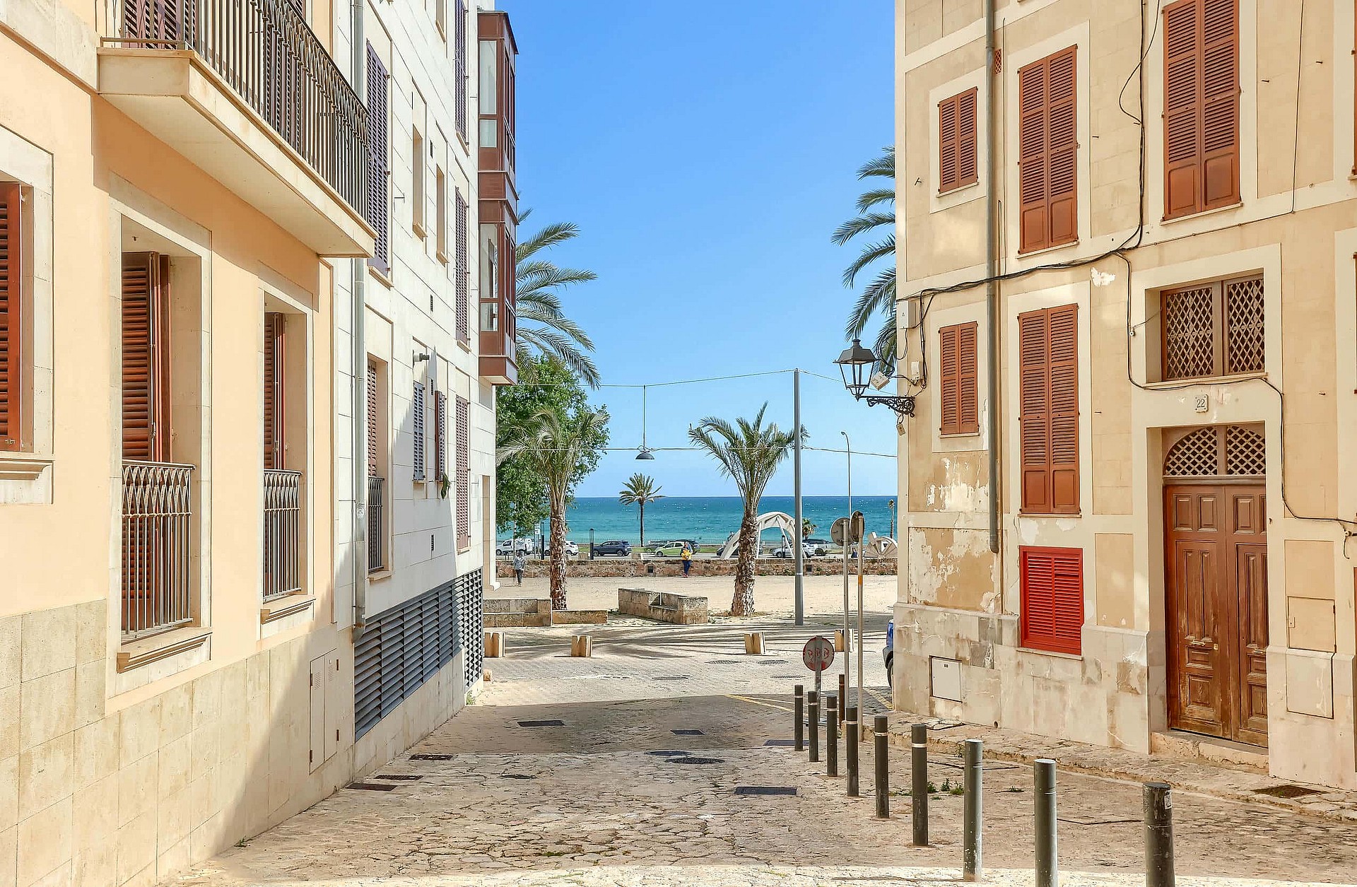 A narrow cobblestone street in a historic Old Town, framed by pale-colored residential buildings with wooden shutters, leading directly to a blue sea view.