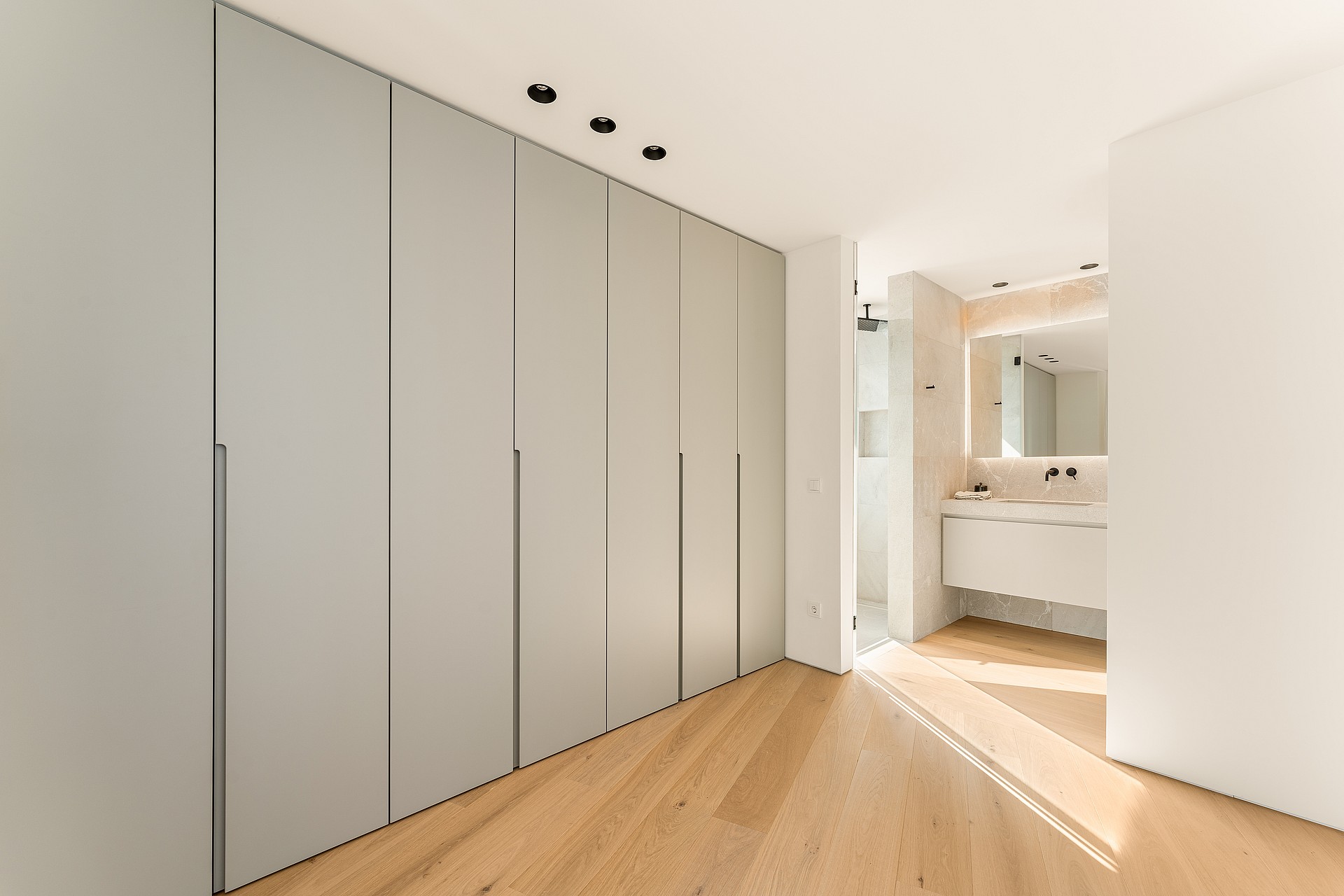 A modern bedroom featuring a floor-to-ceiling grey wardrobe wall, light oak wood flooring, and a view into a bright bathroom.
