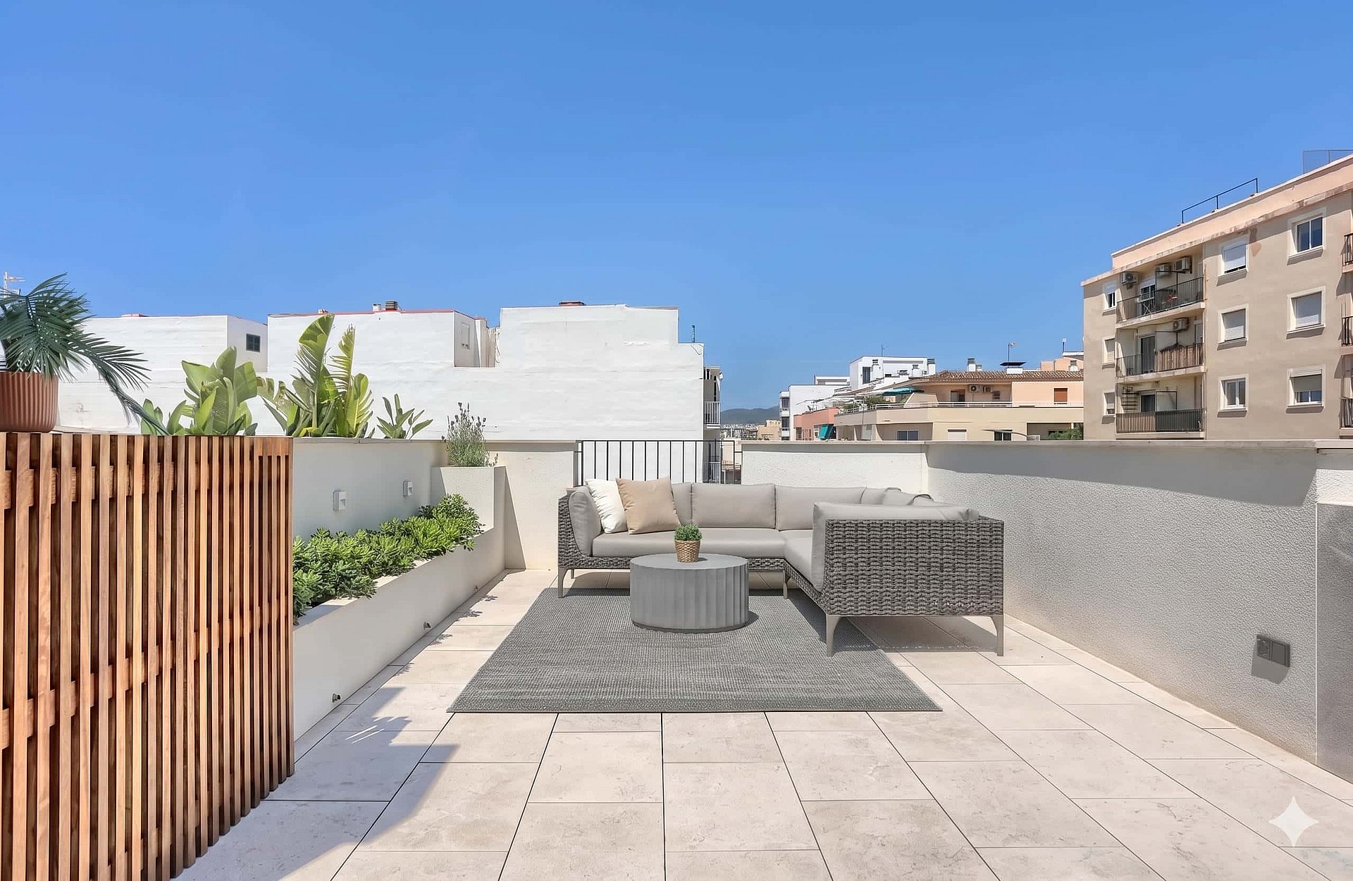 A spacious rooftop terrace featuring a modular outdoor sofa set on a textured rug, bordered by a wooden screen and stone walls.
