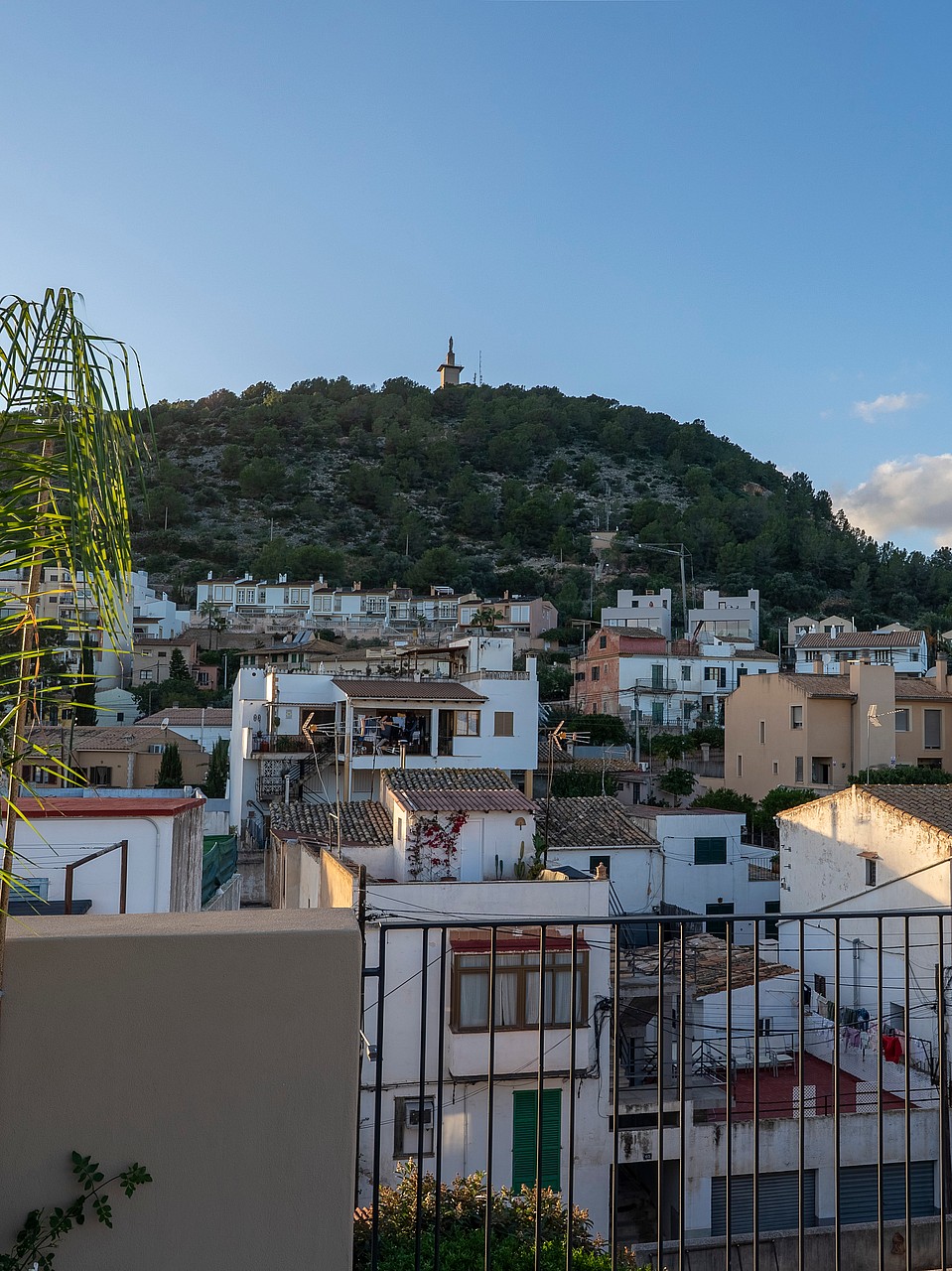 An elevated view across white Mediterranean rooftops toward a green hillside topped with a stone monument.