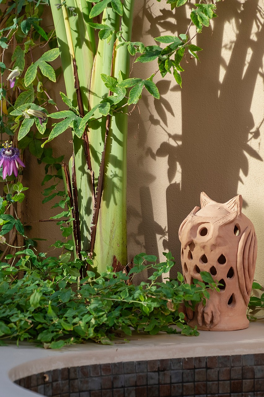 A decorative clay owl sculpture sitting on a stone ledge surrounded by lush green foliage and a tiled pool edge.
