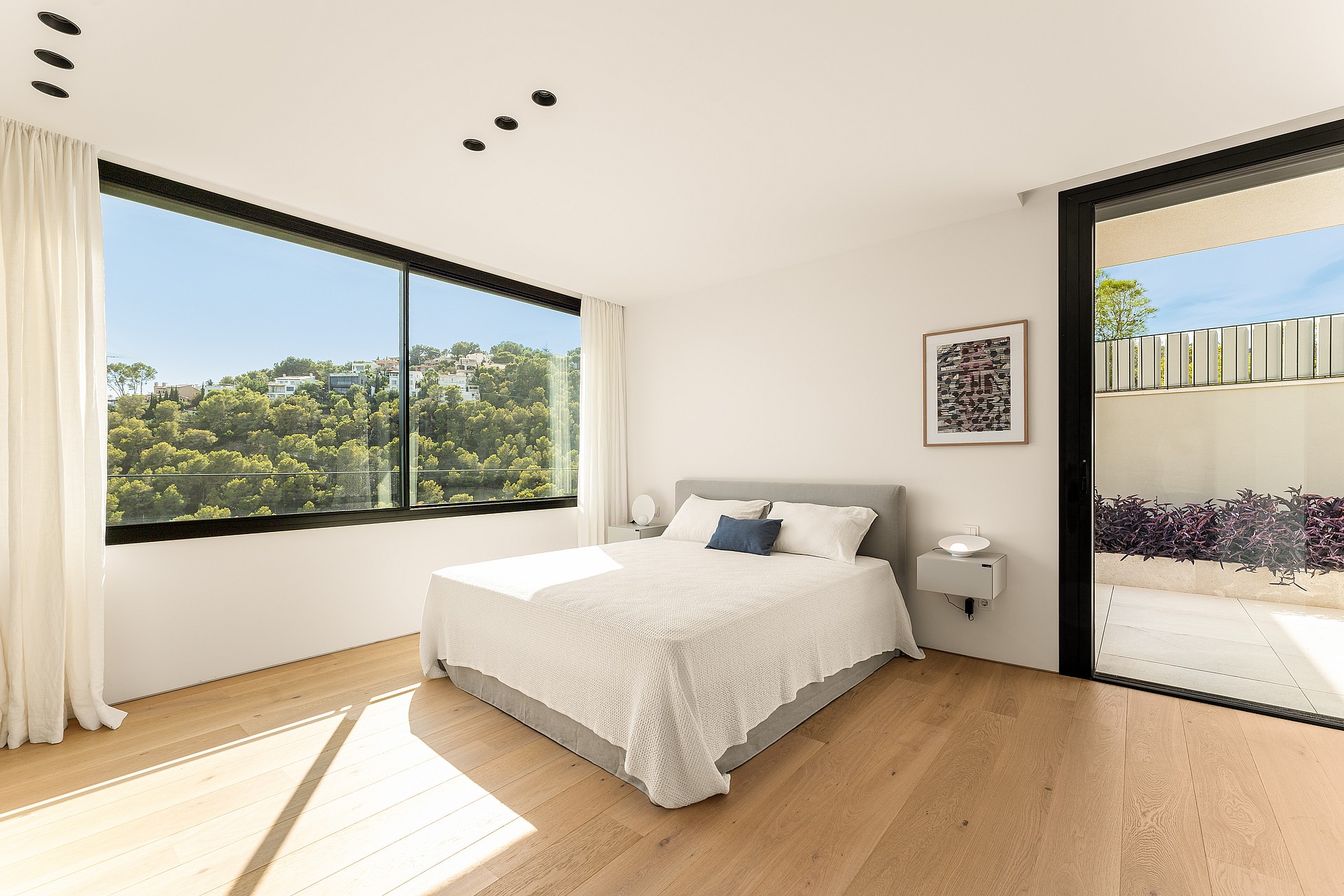 Modern bedroom with oak wood flooring, grey upholstered bed, and floor-to-ceiling windows showing hillside pine trees.