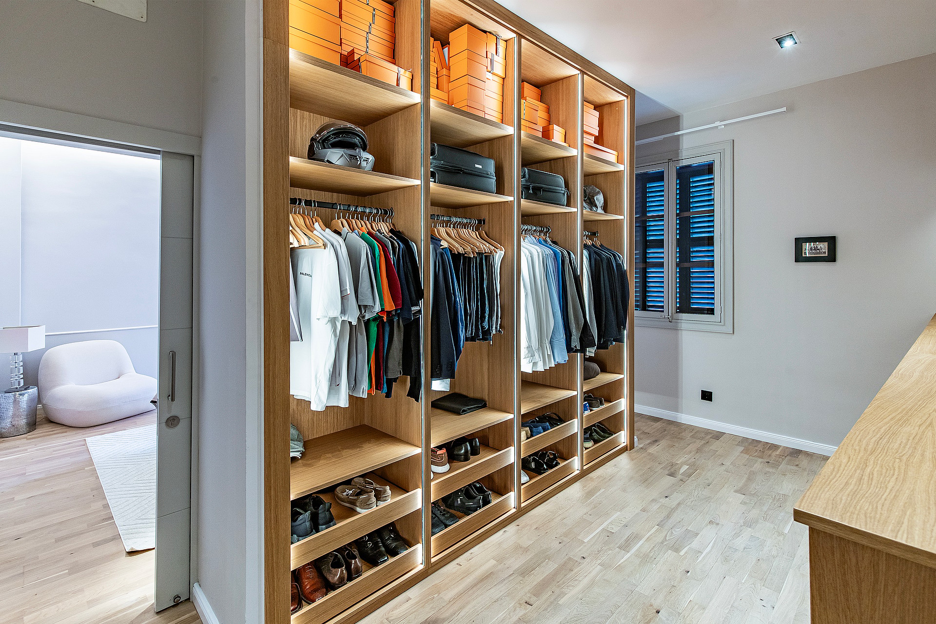 A walk-in dressing room featuring light oak cabinetry with illuminated shelving, hanging rails, and shoe racks, leading to an adjacent bedroom.