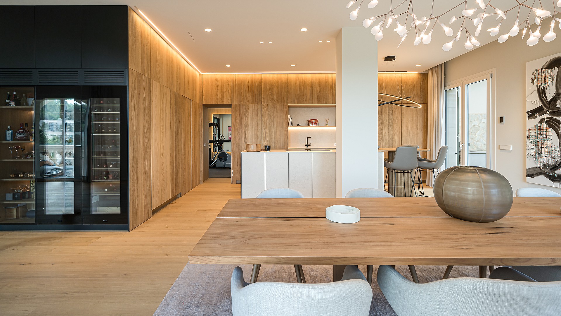 Dining room featuring a long wooden table, light grey upholstered chairs, and a floor-to-ceiling built-in wine fridge in a black frame.