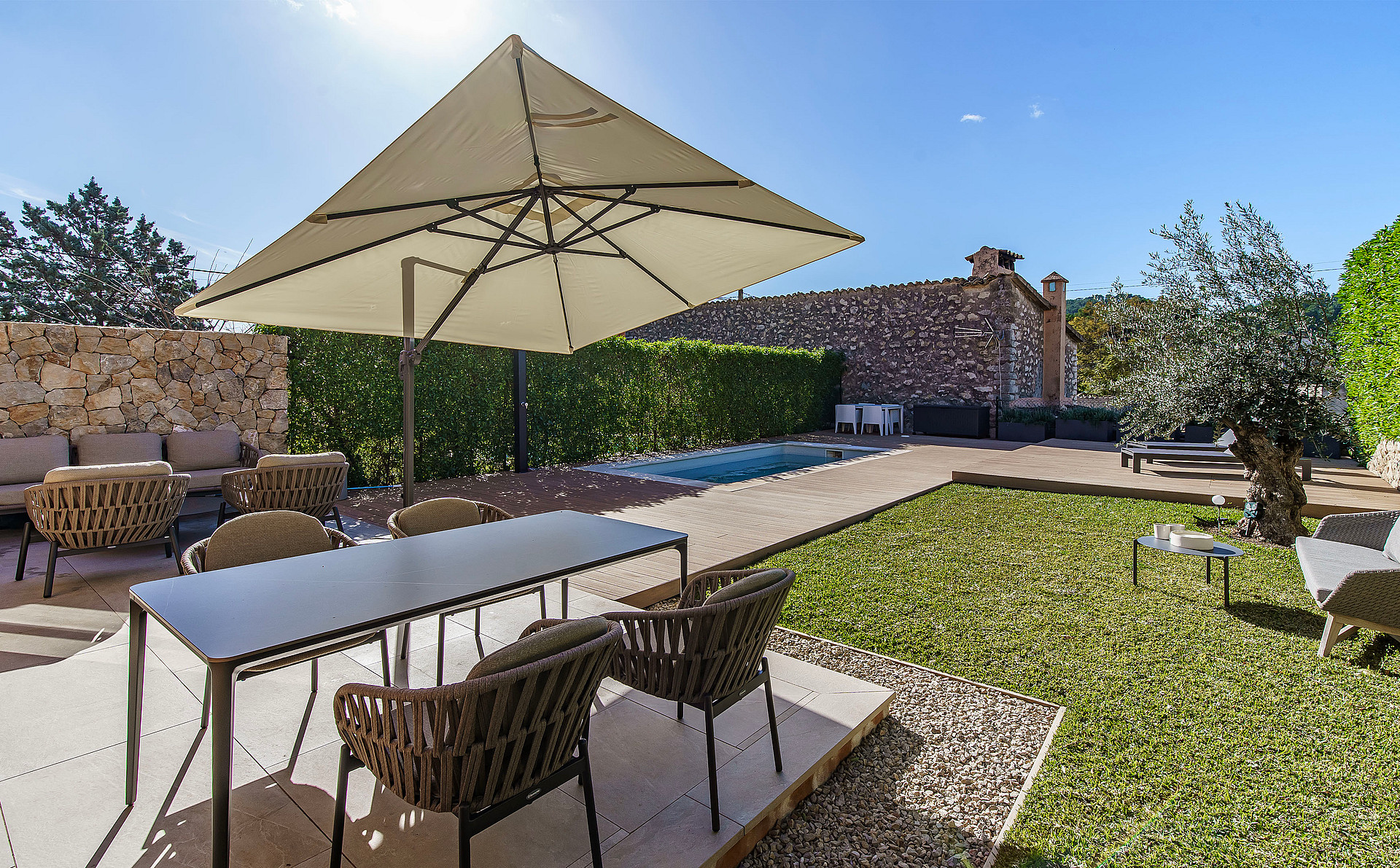 An outdoor dining terrace in Es Capdella featuring a rectangular table with woven chairs, a large beige parasol, and a small pool set against a natural stone wall.