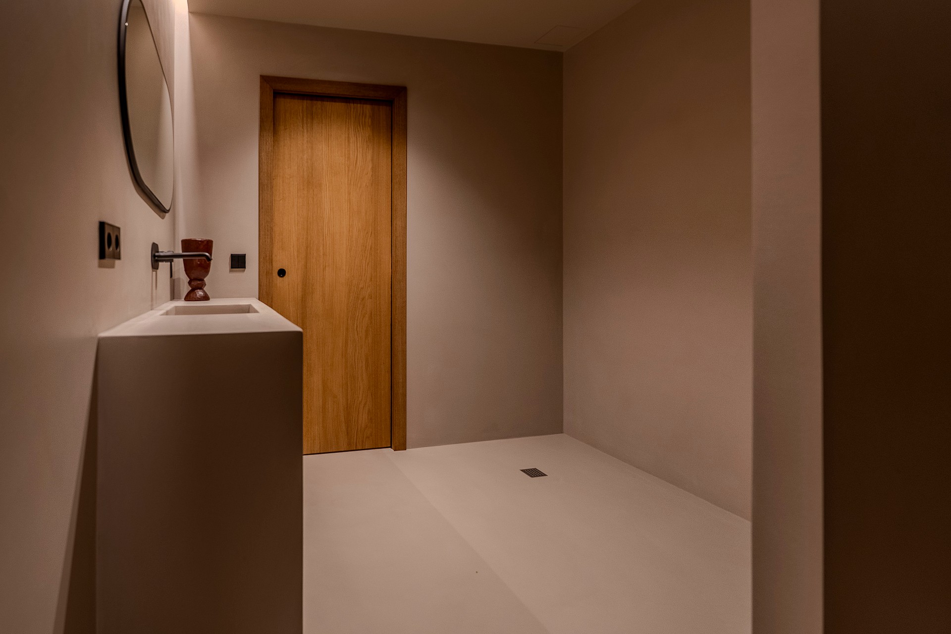 A minimalist bathroom featuring a white monolith vanity, a circular mirror, and a natural wood door under warm lighting.