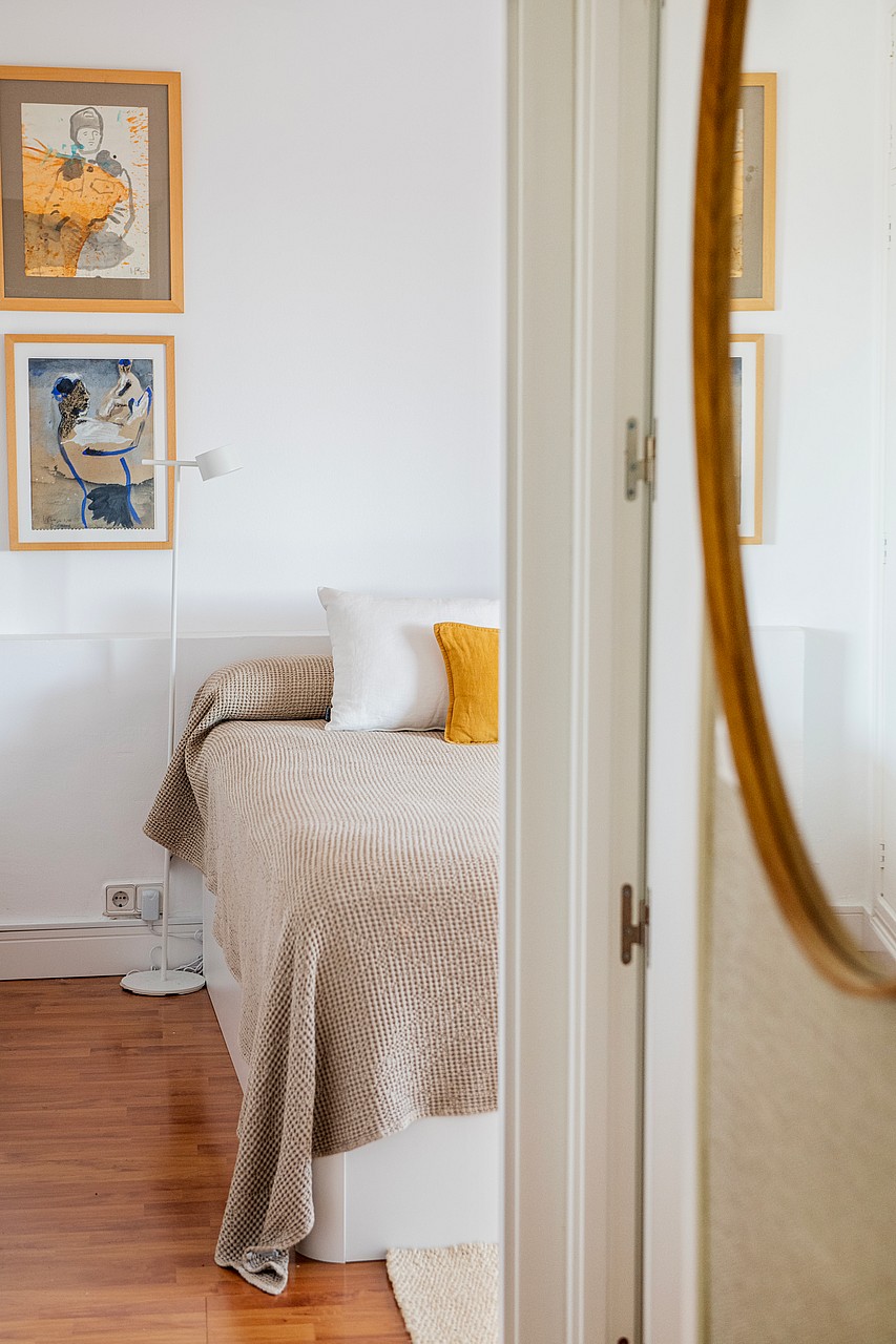 A close-up view of a bedroom interior with a bed featuring textured bedding, two framed artworks on the white wall, and a large oval mirror in the foreground.