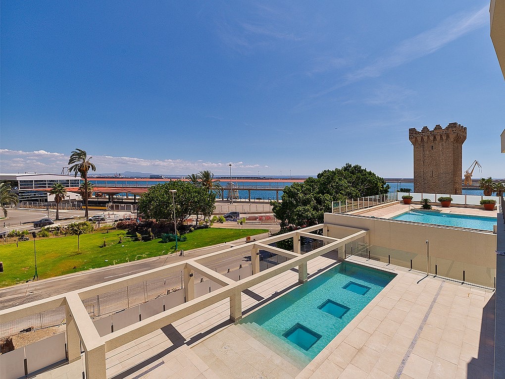 Vista en ángulo de una terraza con piscina rectangular, sobre un parque, la carretera costera y el mar.