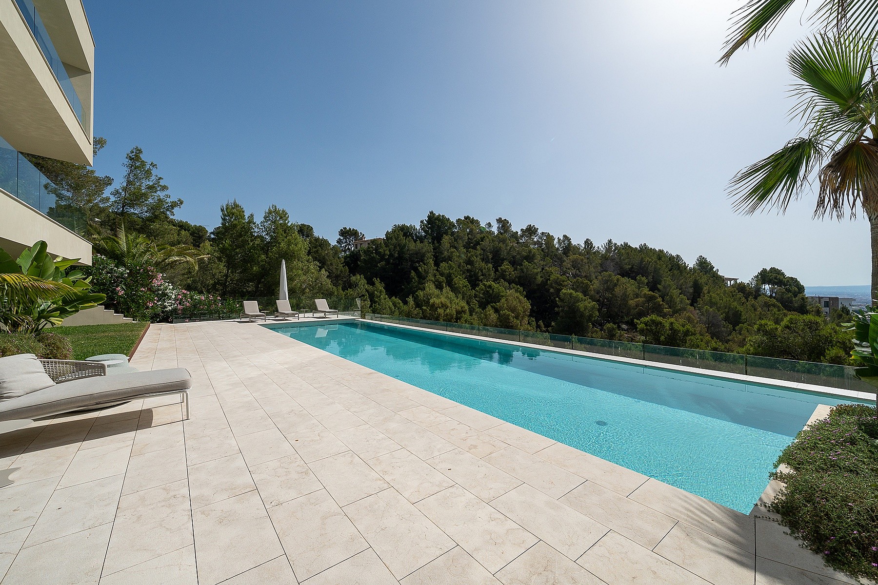 A long rectangular swimming pool with a stone-paved deck, lounge chairs, and a glass balustrade overlooking a tree-lined hill.