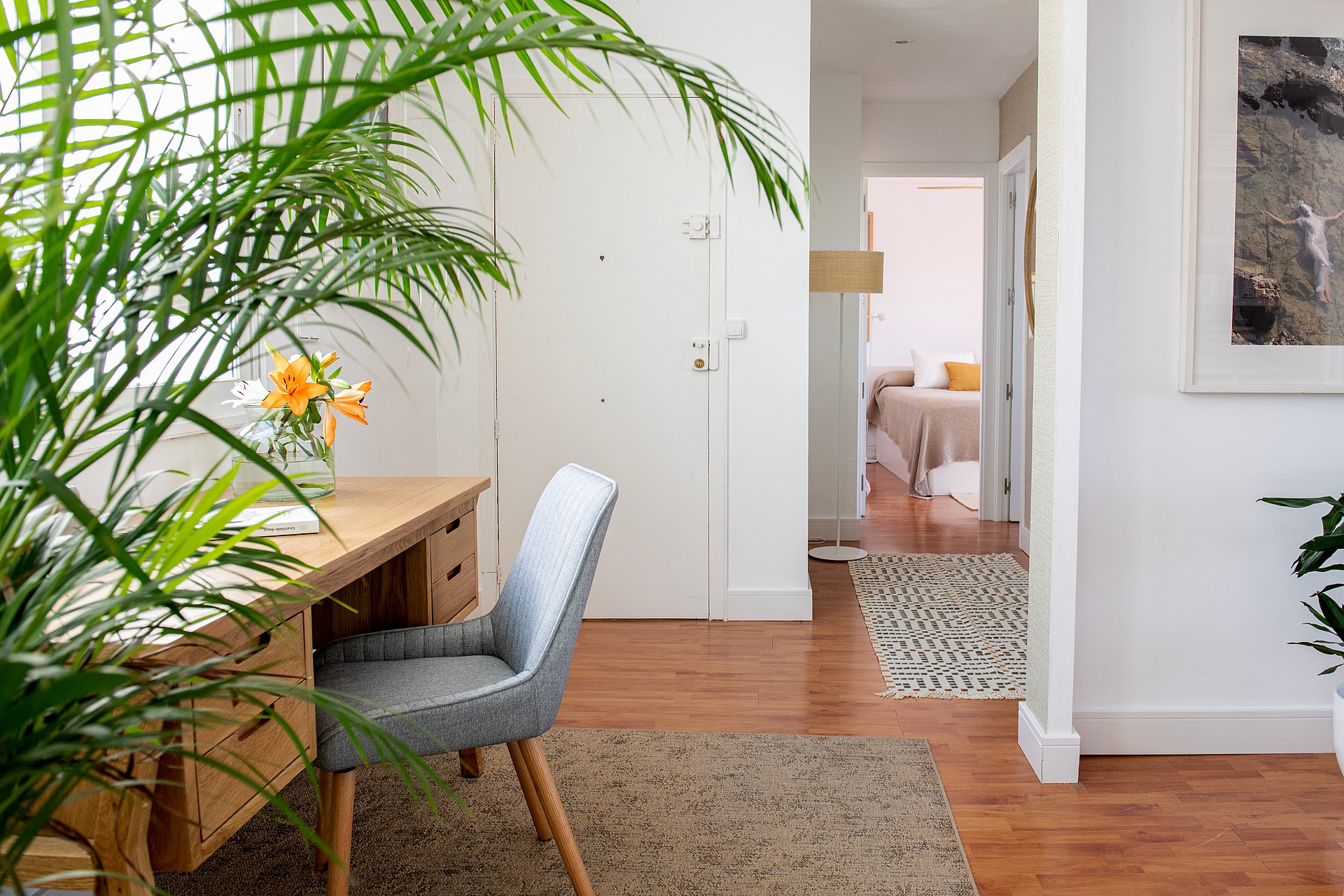 A home office area with a light wood desk, grey upholstered chair, and potted plant in the foreground, leading to a bedroom.