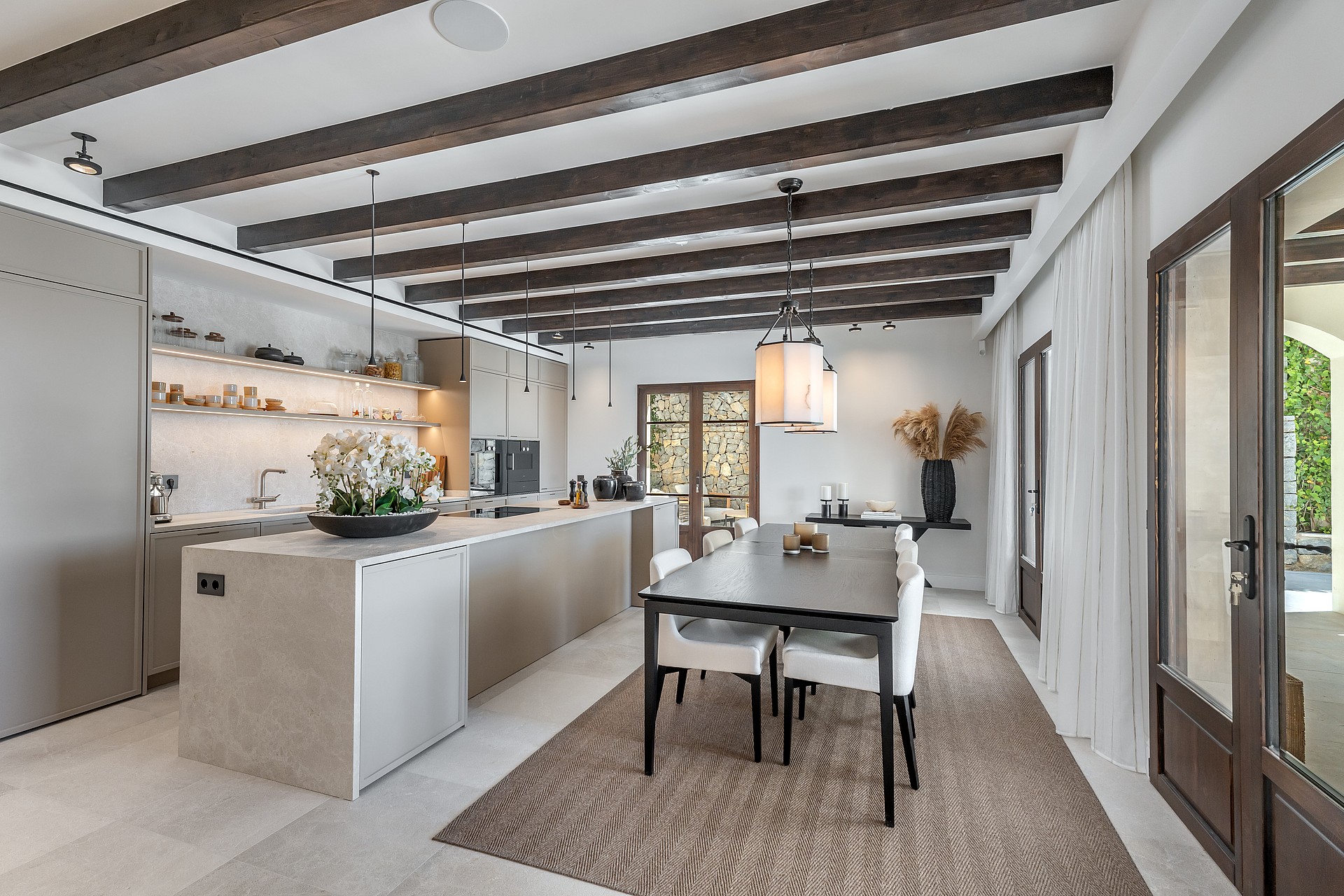 Modern dining area showing dark wood ceiling beams, a central table with white chairs, and an adjacent kitchen island.