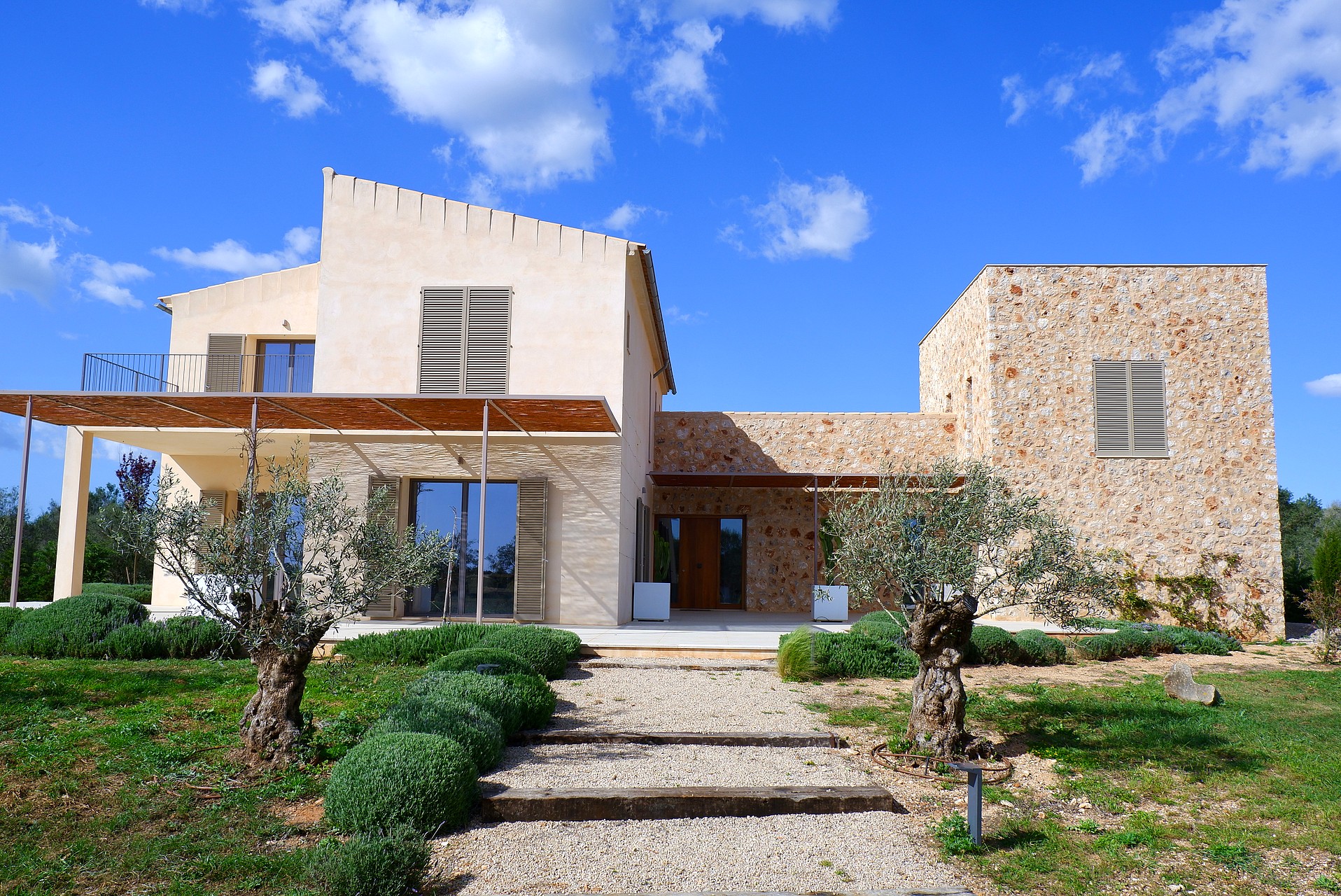 Two-story stone-clad finca with a graveled pathway, mature olive trees, and wooden pergola under a clear blue sky.