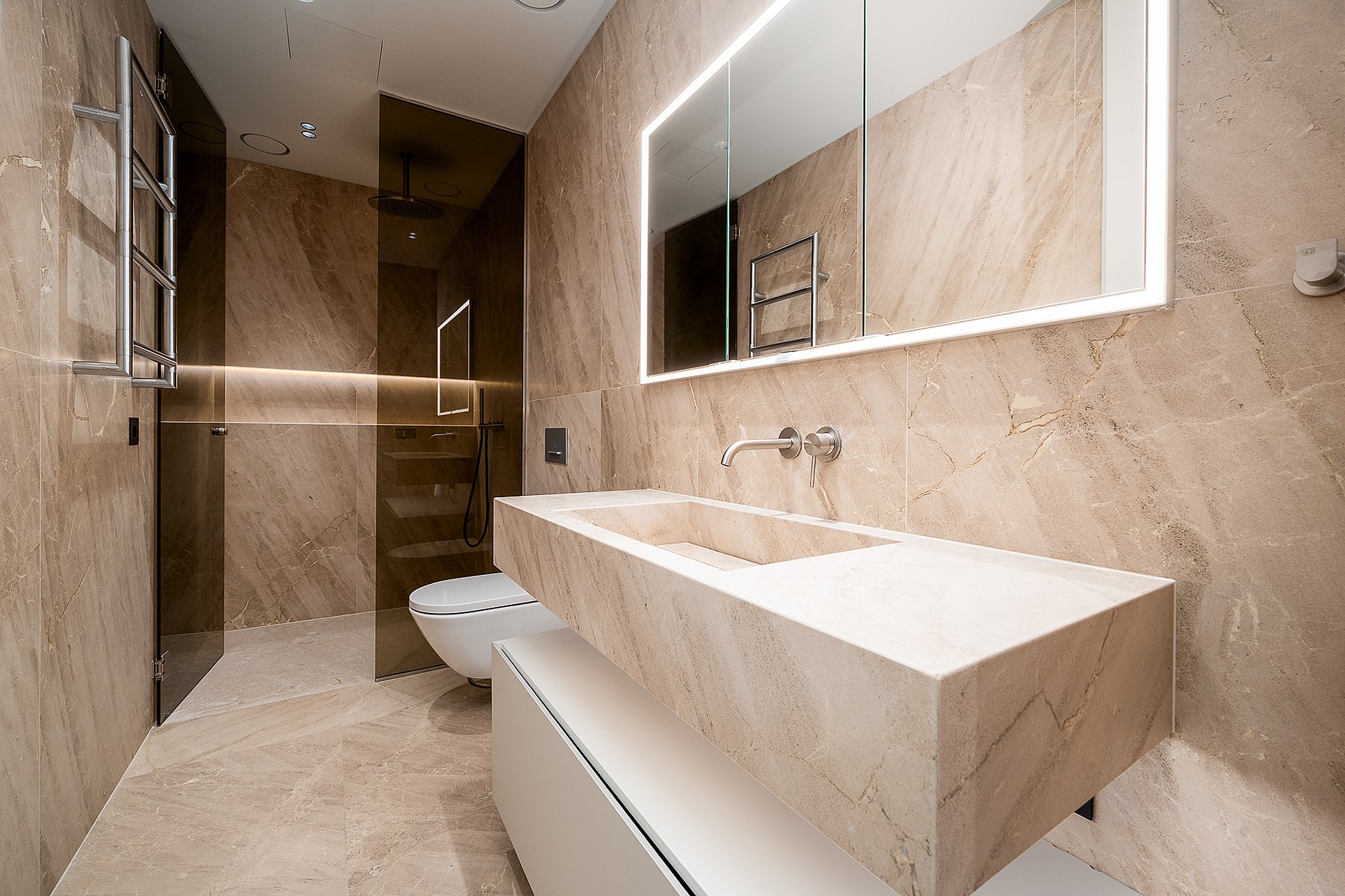Modern bathroom featuring beige stone wall cladding, a floating stone vanity sink, a wall-mounted toilet, and a glass-enclosed shower area with integrated LED mirror lighting.
