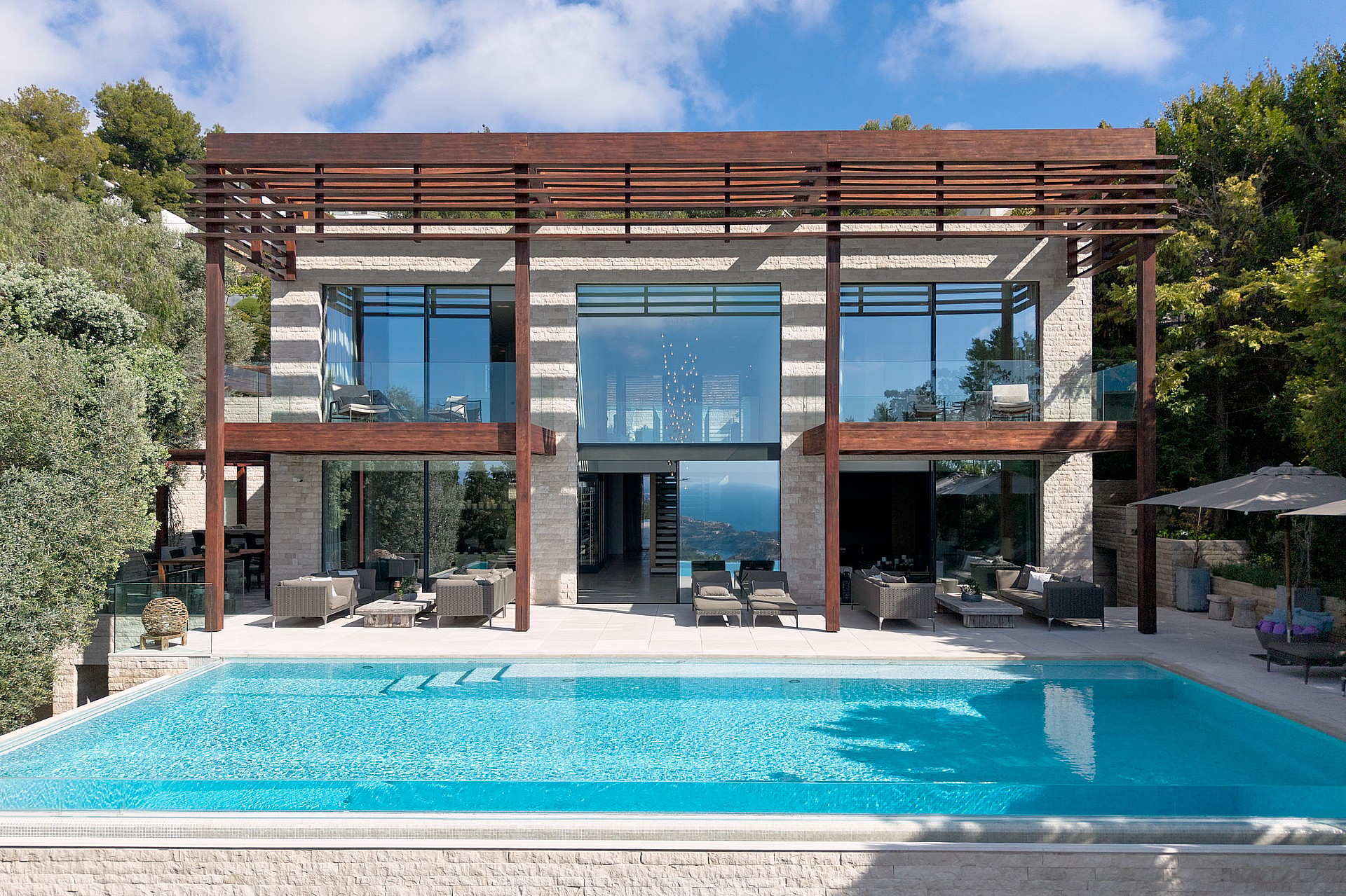 Exterior of a contemporary villa featuring an infinity pool, natural stone facade, floor-to-ceiling glass, and timber pergola.