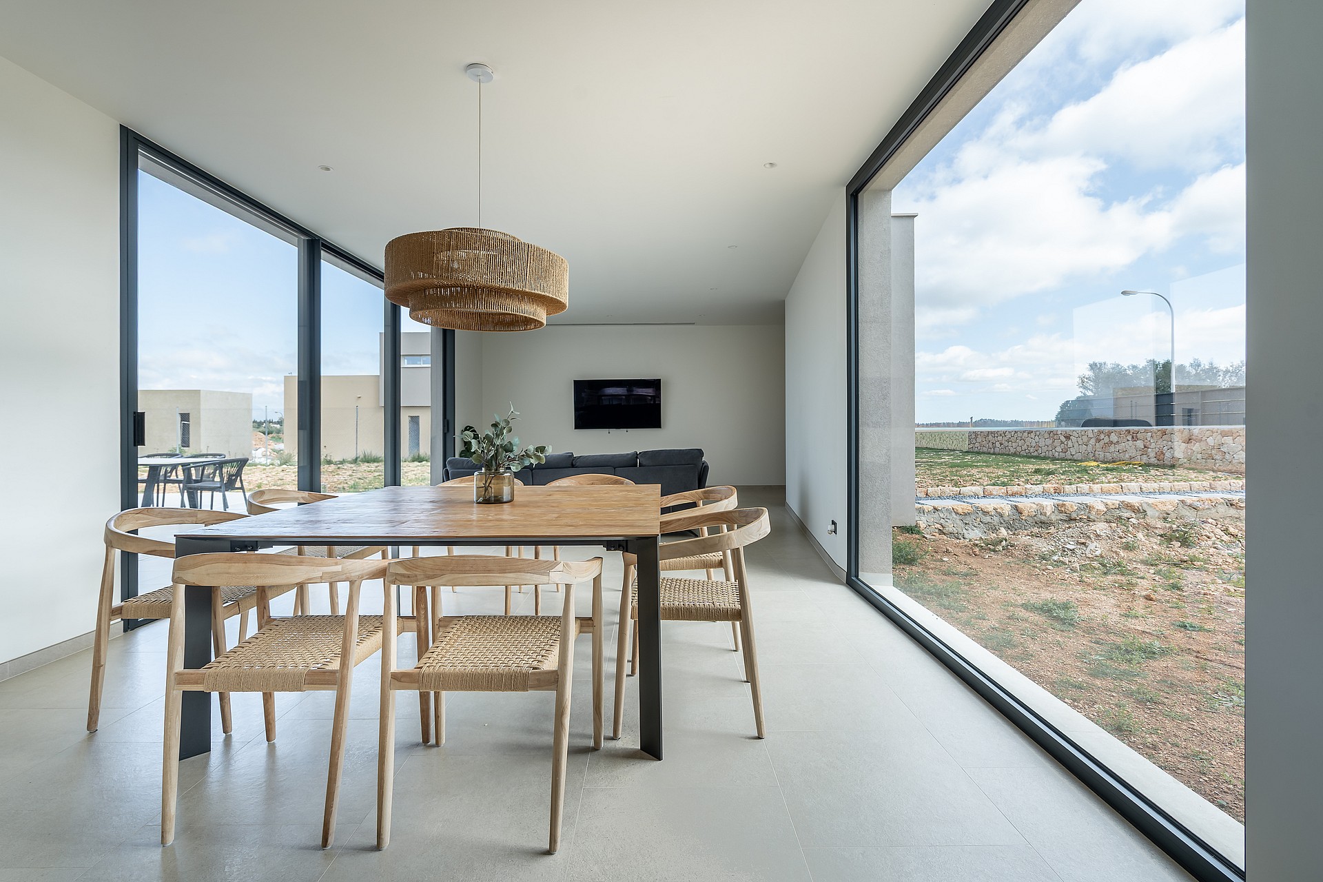 Dining room with a wooden table, woven chairs, and large glass windows overlooking a dry landscape.