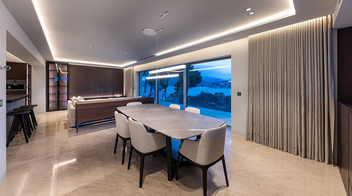 Modern dining room with stone table, upholstered chairs, and floor-to-ceiling glass windows overlooking a coastal landscape.