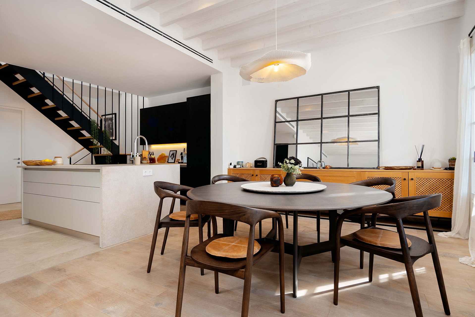 Dining room with a round wooden table, black chairs, and a metal staircase leading to the upper floor.