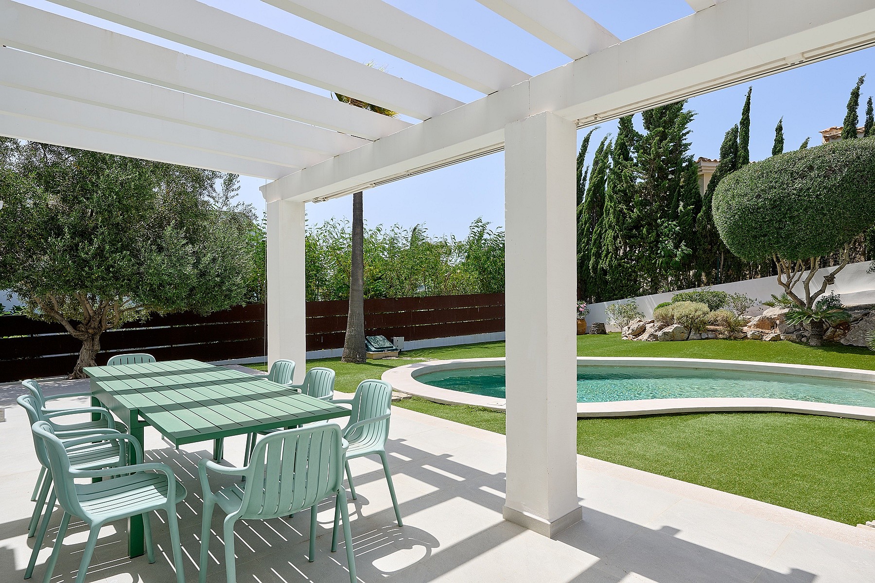 Pérgola blanca sobre mesa de comedor exterior junto a piscina circular con cipreses y setos perfilados