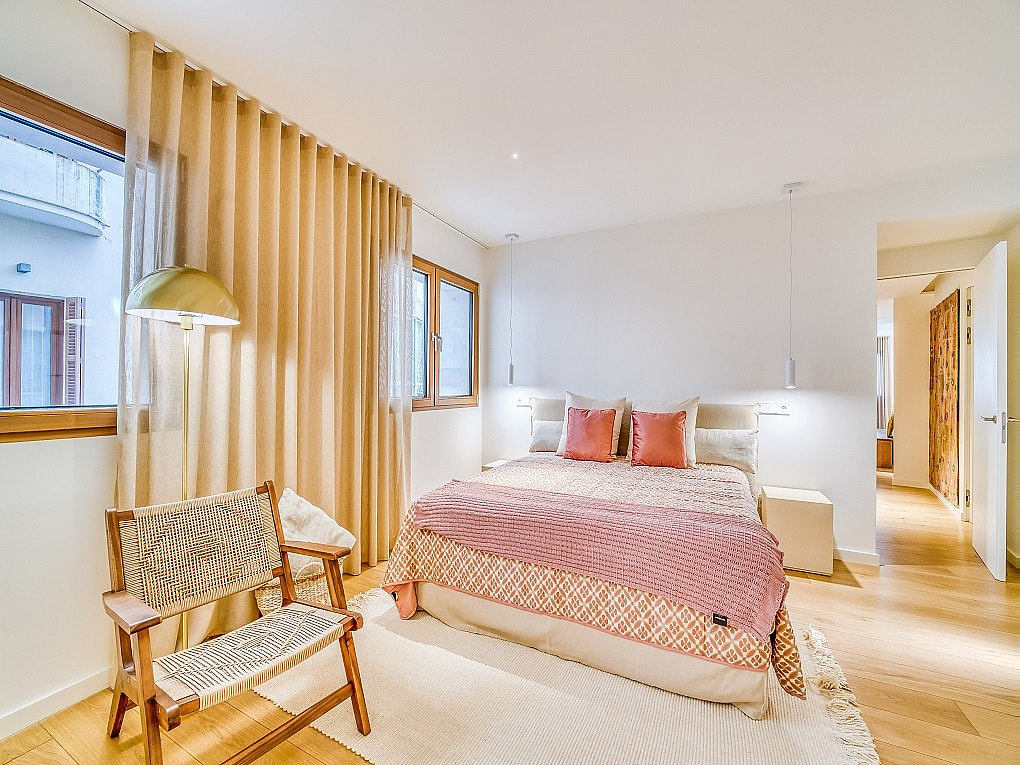 A bright bedroom with light wooden floors, a double bed with coral bedding, and a wooden armchair beside floor-length curtains.