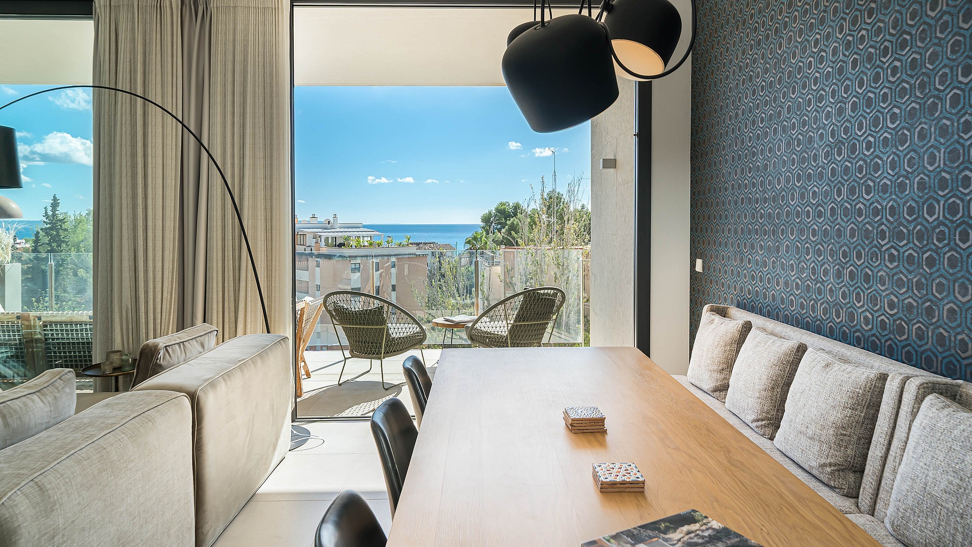 Dining room with wooden table, modern chairs, and black pendant lights, opening to a terrace with wicker armchairs and sea views.