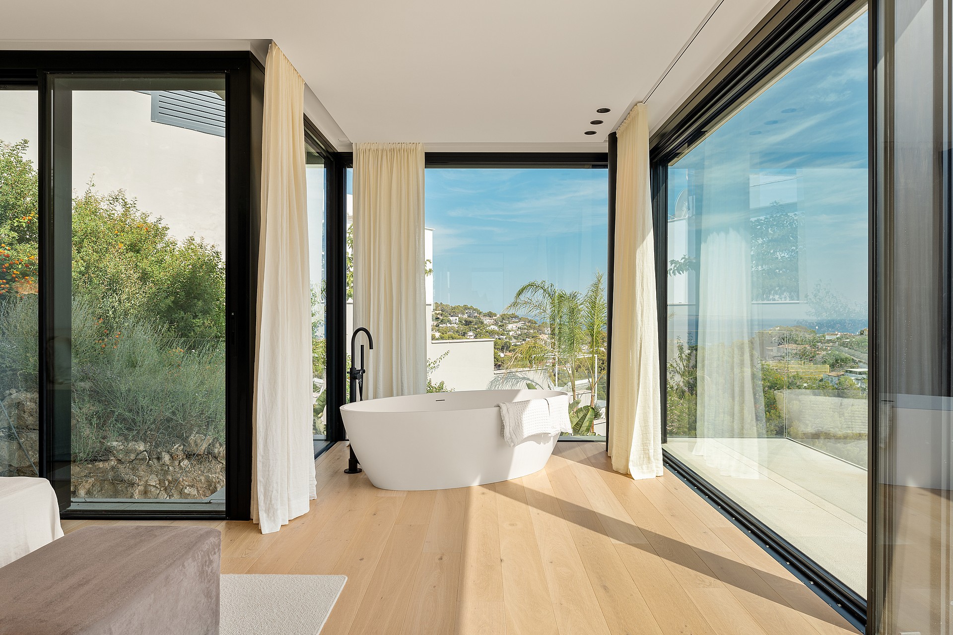 A modern master bathroom features a white freestanding oval bathtub, light oak hardwood floors, and floor-to-ceiling black-framed windows overlooking a lush garden.