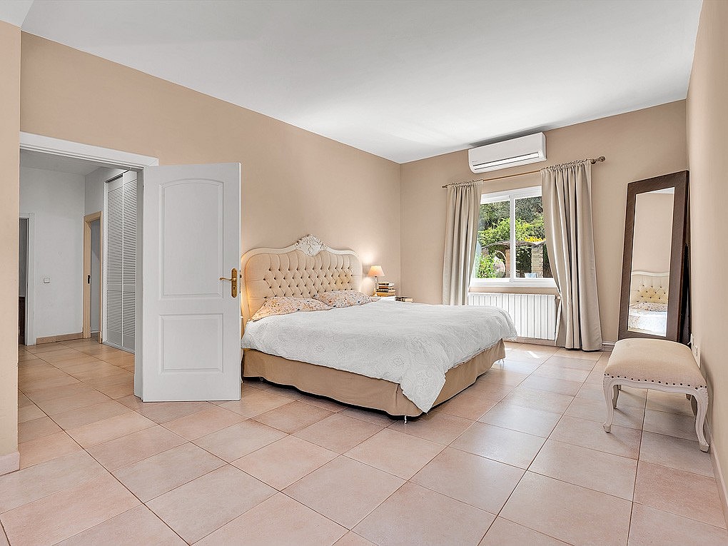 A spacious bedroom featuring a tufted upholstered bed, light beige walls, tiled floors, and a floor mirror.