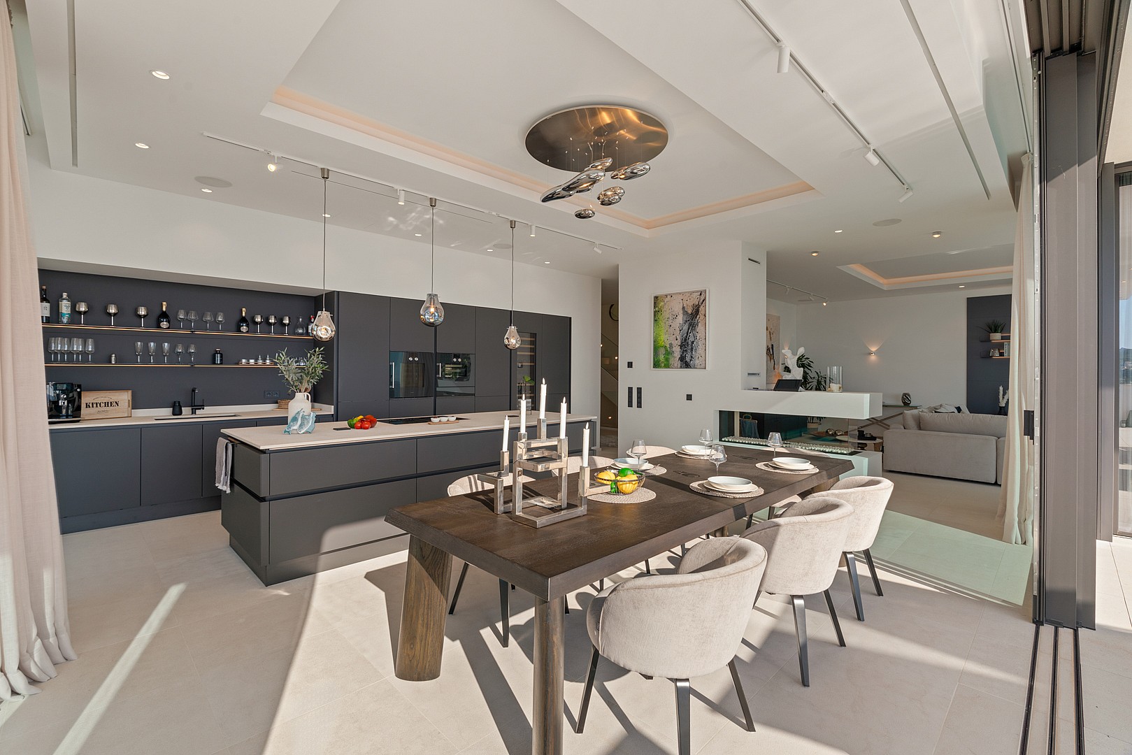 Modern open-concept interior featuring a dark grey kitchen island, a wooden dining table with light chairs, and recessed ceiling lighting.