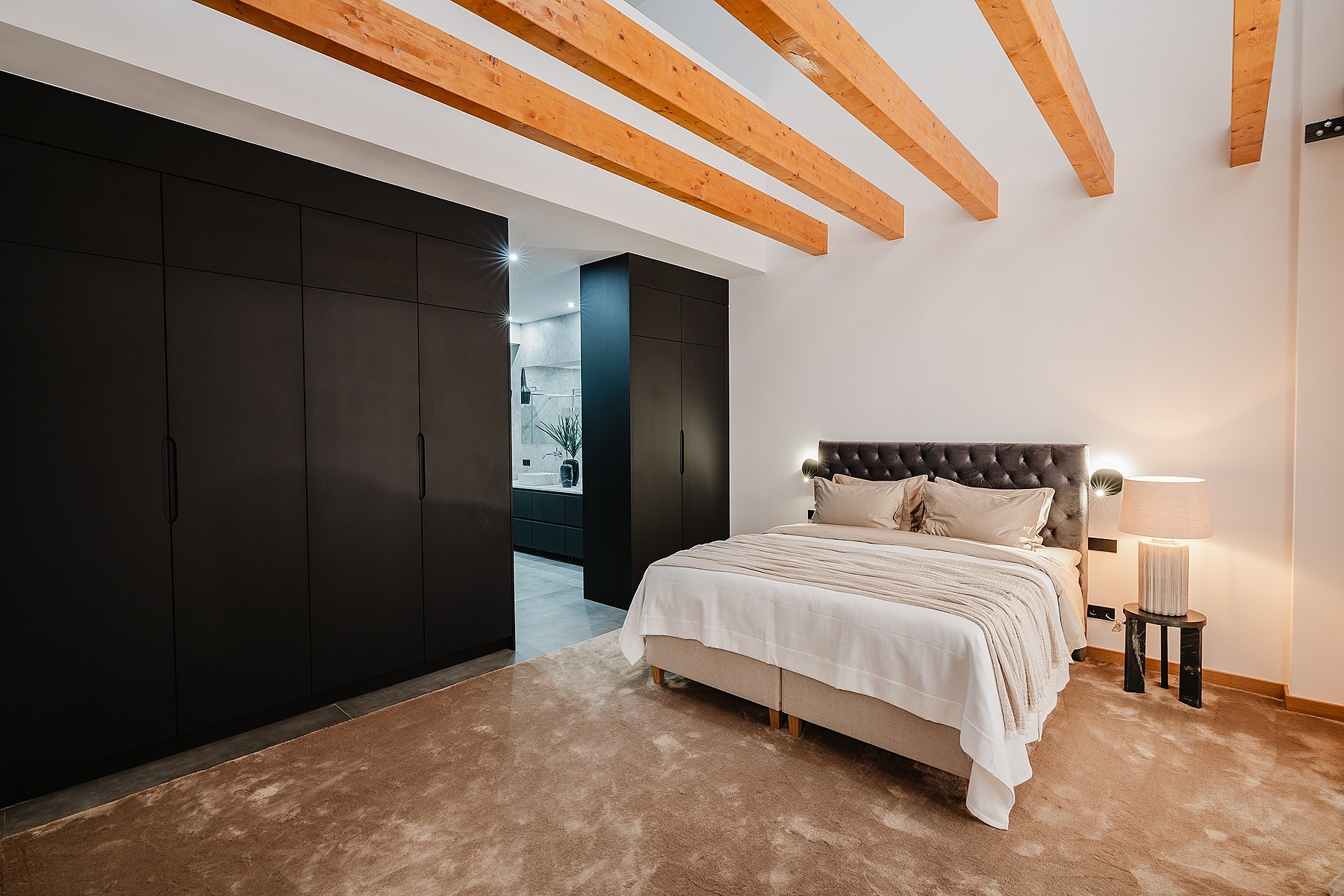 A modern bedroom with a large bed against a white wall, dark floor-to-ceiling wardrobes, and visible natural wood ceiling beams.