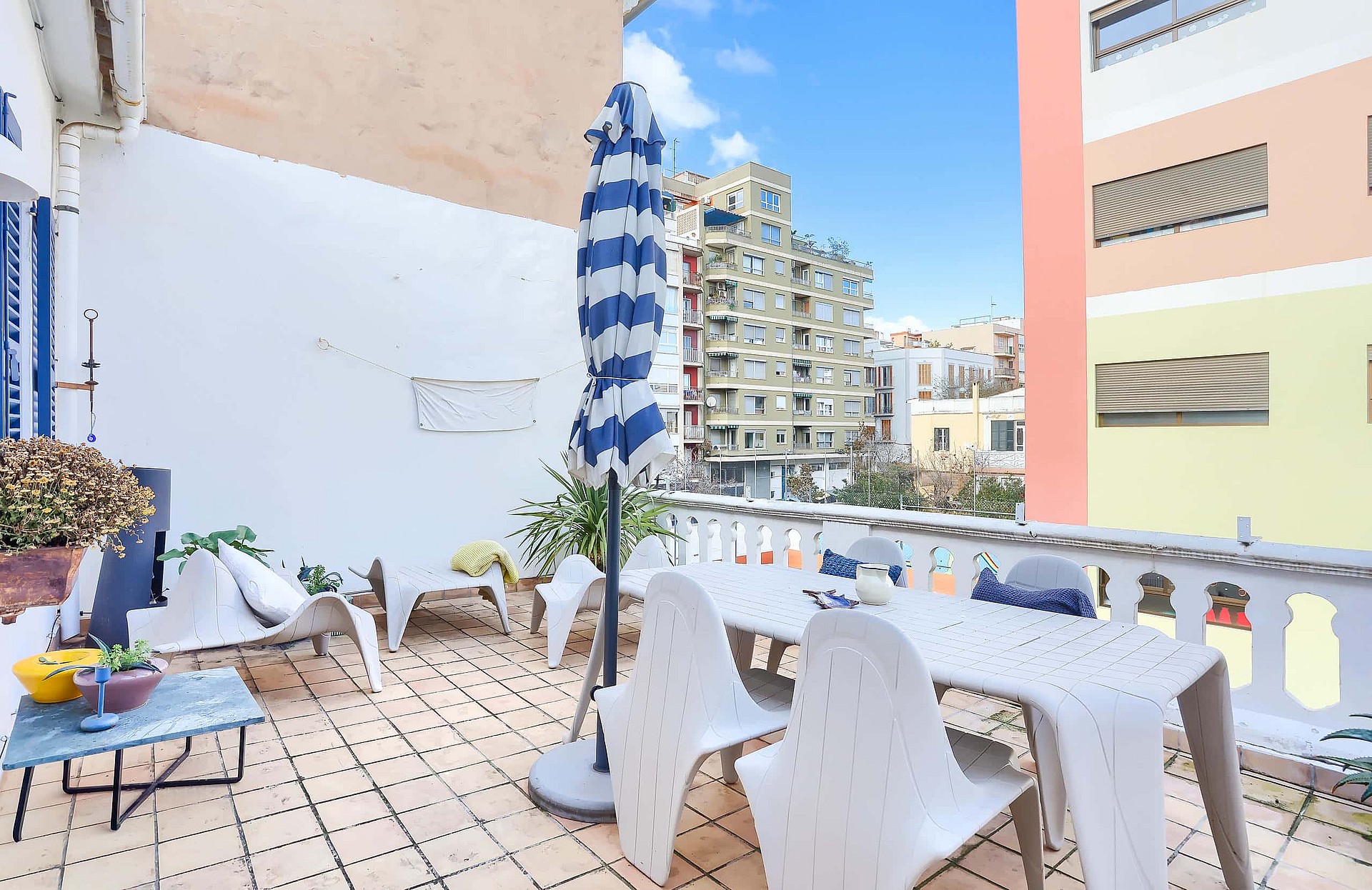 A sunny outdoor terrace with a dining table, chairs, and a blue-striped parasol, featuring city views of neighboring buildings.