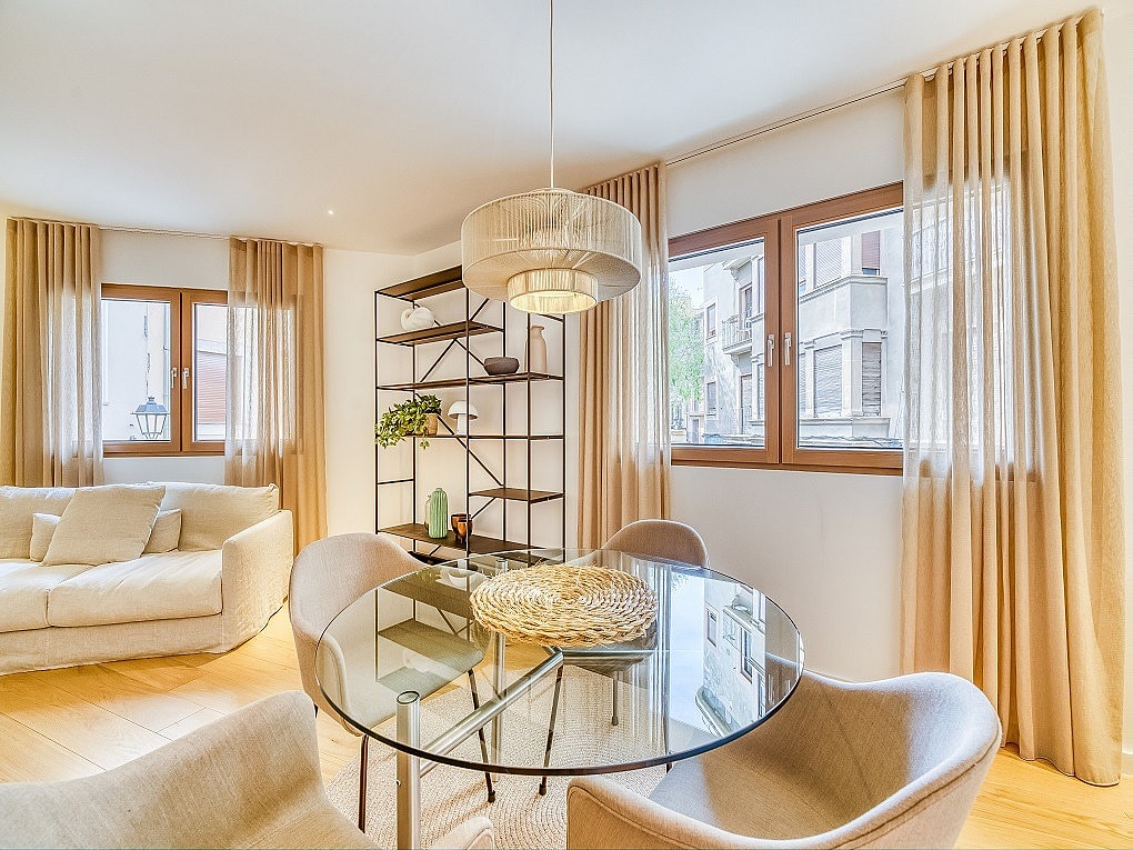 A glass dining table with four curved chairs next to a metal shelving unit in an apartment, with linen curtains framing city windows.
