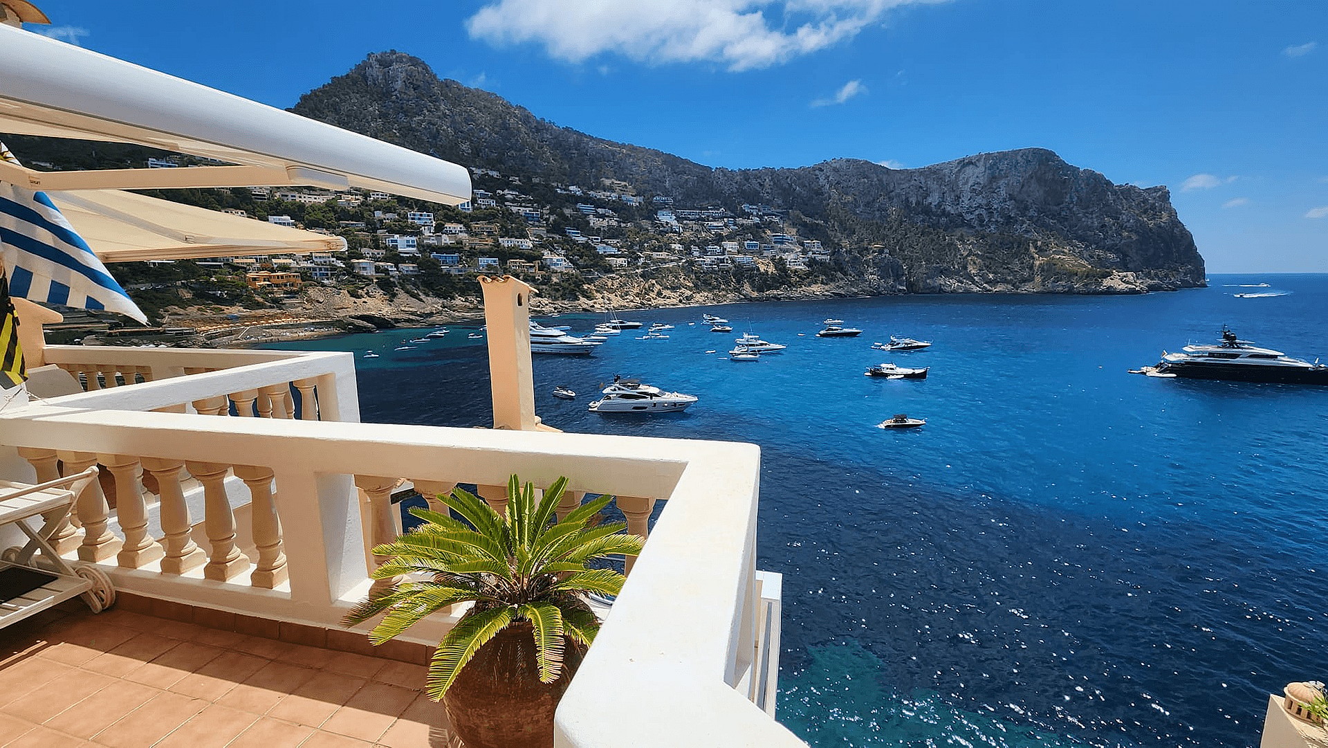 A wide-angle shot of a terrace with white balustrades overlooking the blue waters of Cala Llamp, featuring mountains and anchored boats.