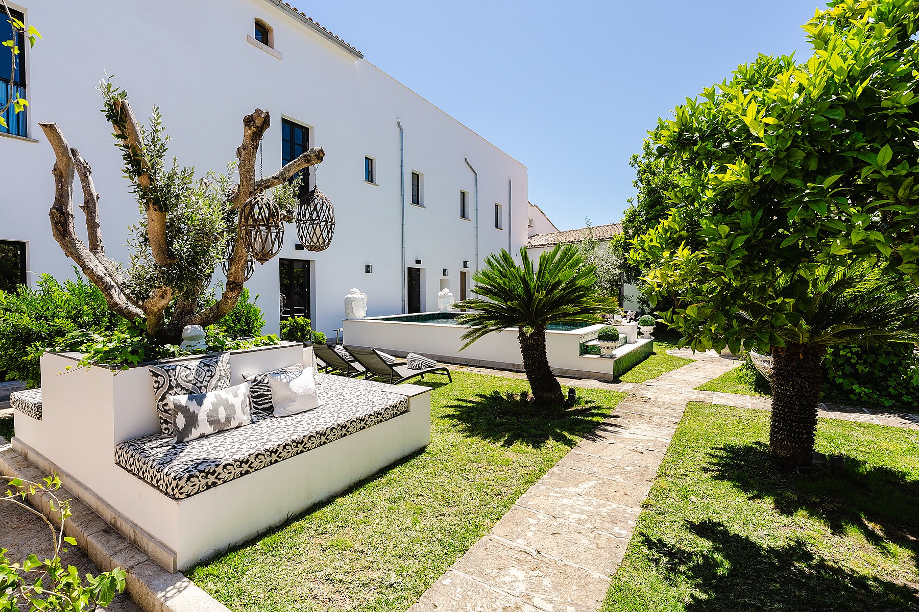 A white townhouse exterior with a landscaped garden featuring a built-in stone bench, patterned cushions, and a palm tree.