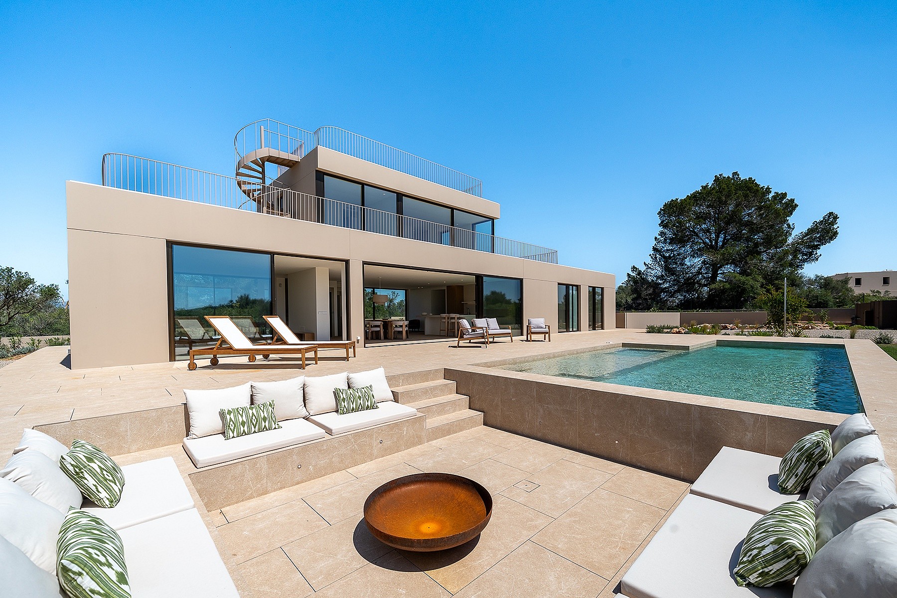 A modern villa exterior featuring a sunken stone lounge with built-in seating, a circular fire pit, and an adjacent swimming pool under a clear blue sky.