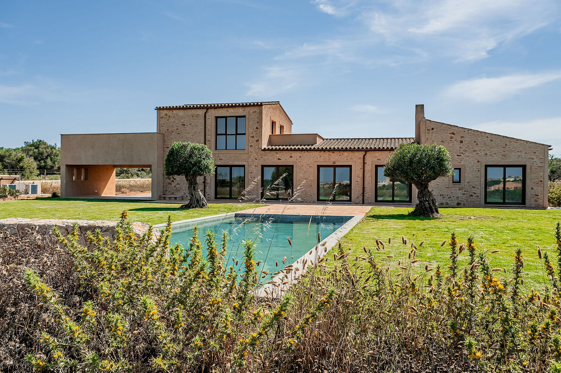 Exterior view of a traditional stone finca with a swimming pool, manicured lawn, and native trees under a clear blue sky.