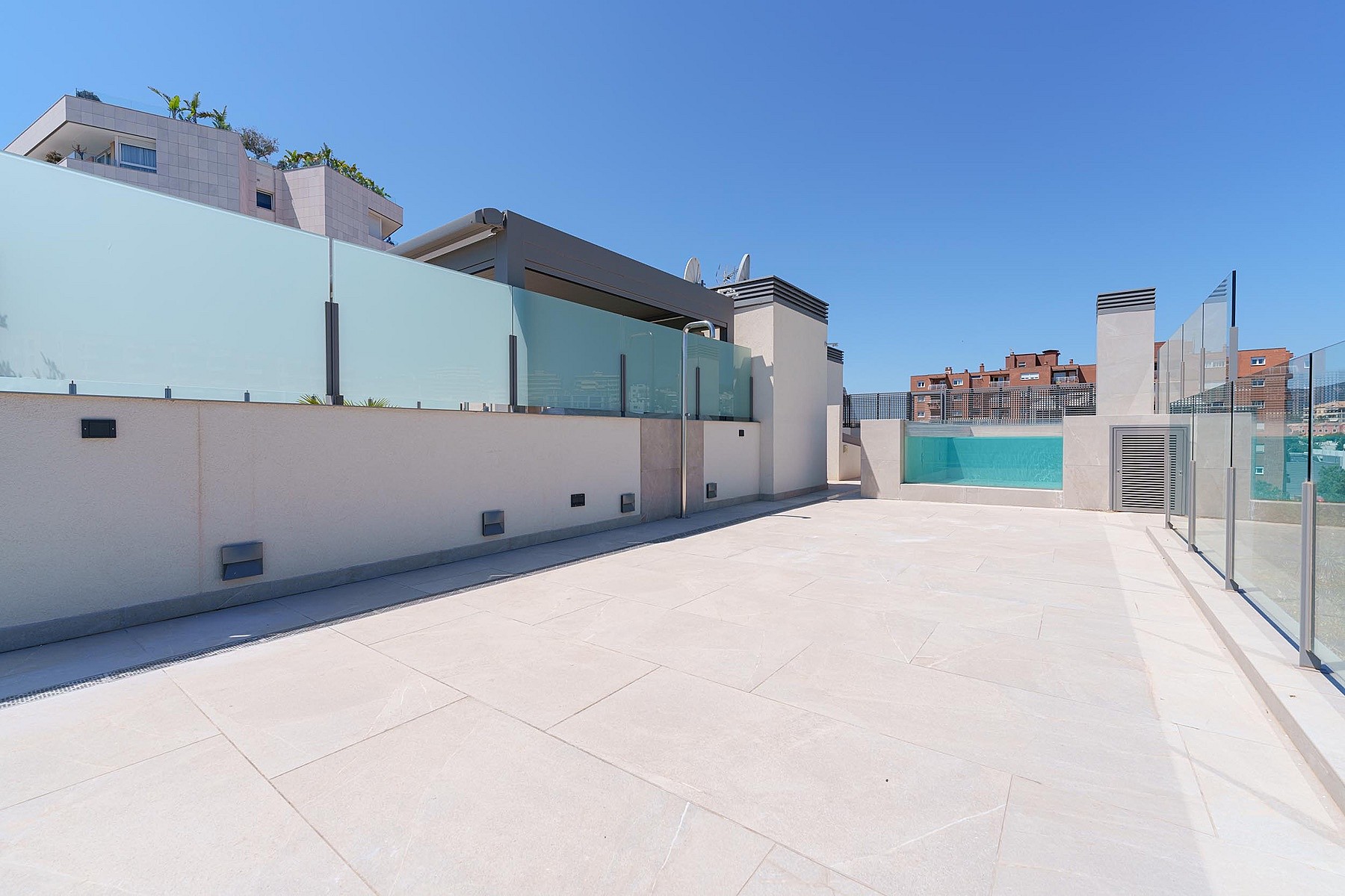 Terraza espaciosa en azotea con baldosas de piedra, delimitada por paneles de cristal bajo un cielo azul.