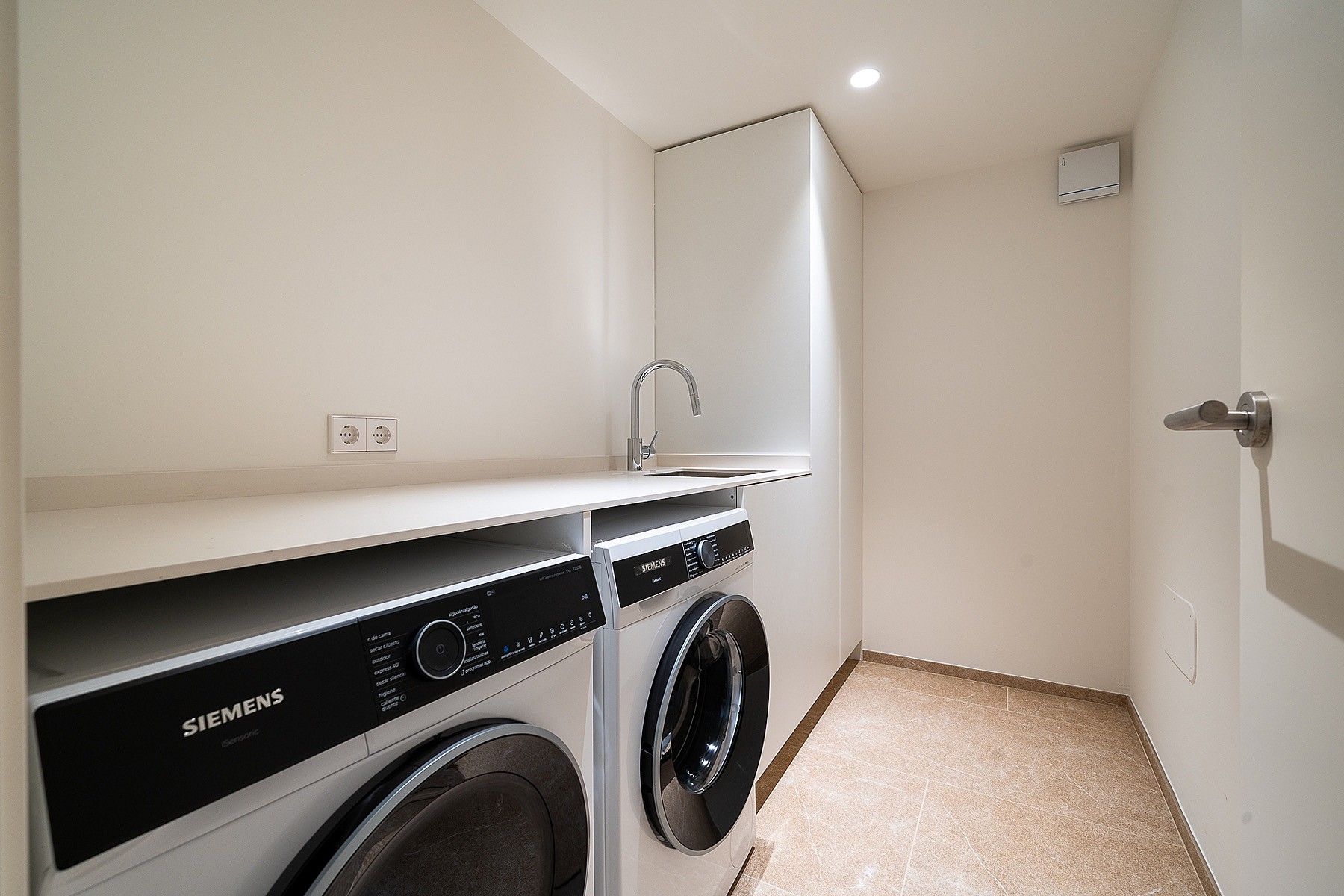 A white minimalist laundry room featuring a stacked washer and dryer, white countertops, a small steel sink, and light flooring.