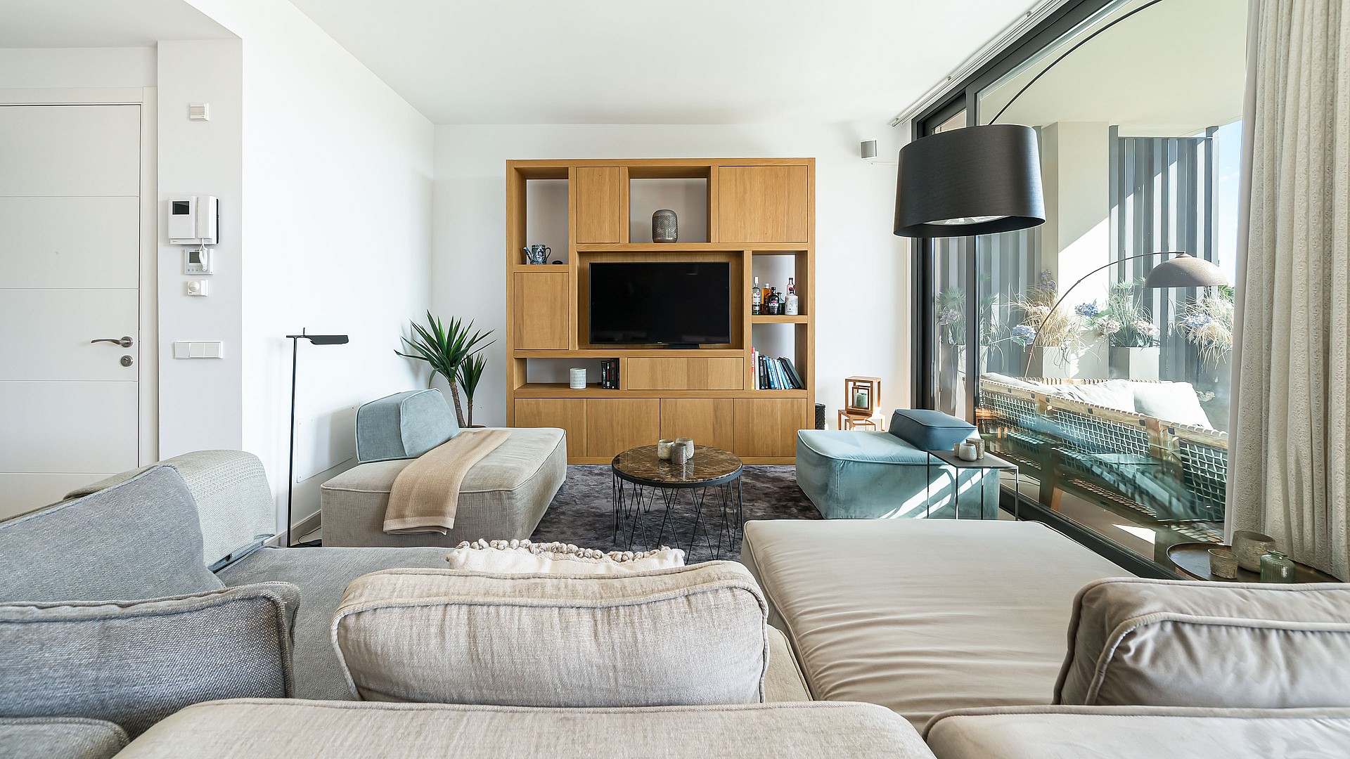 Modern living room with a large modular sofa, oak wood media wall unit, and glass sliding doors overlooking a terrace.