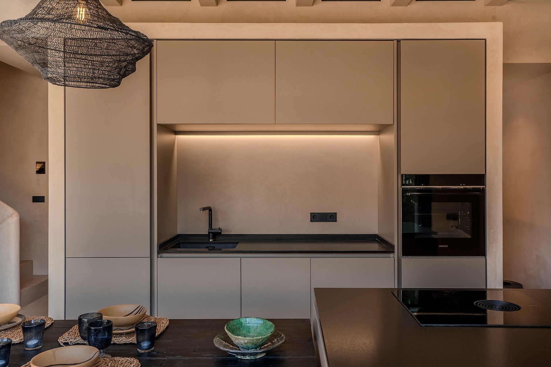 A minimalist kitchen wall featuring matte beige cabinetry, a dark countertop, integrated oven, and a sculptural textured pendant light.