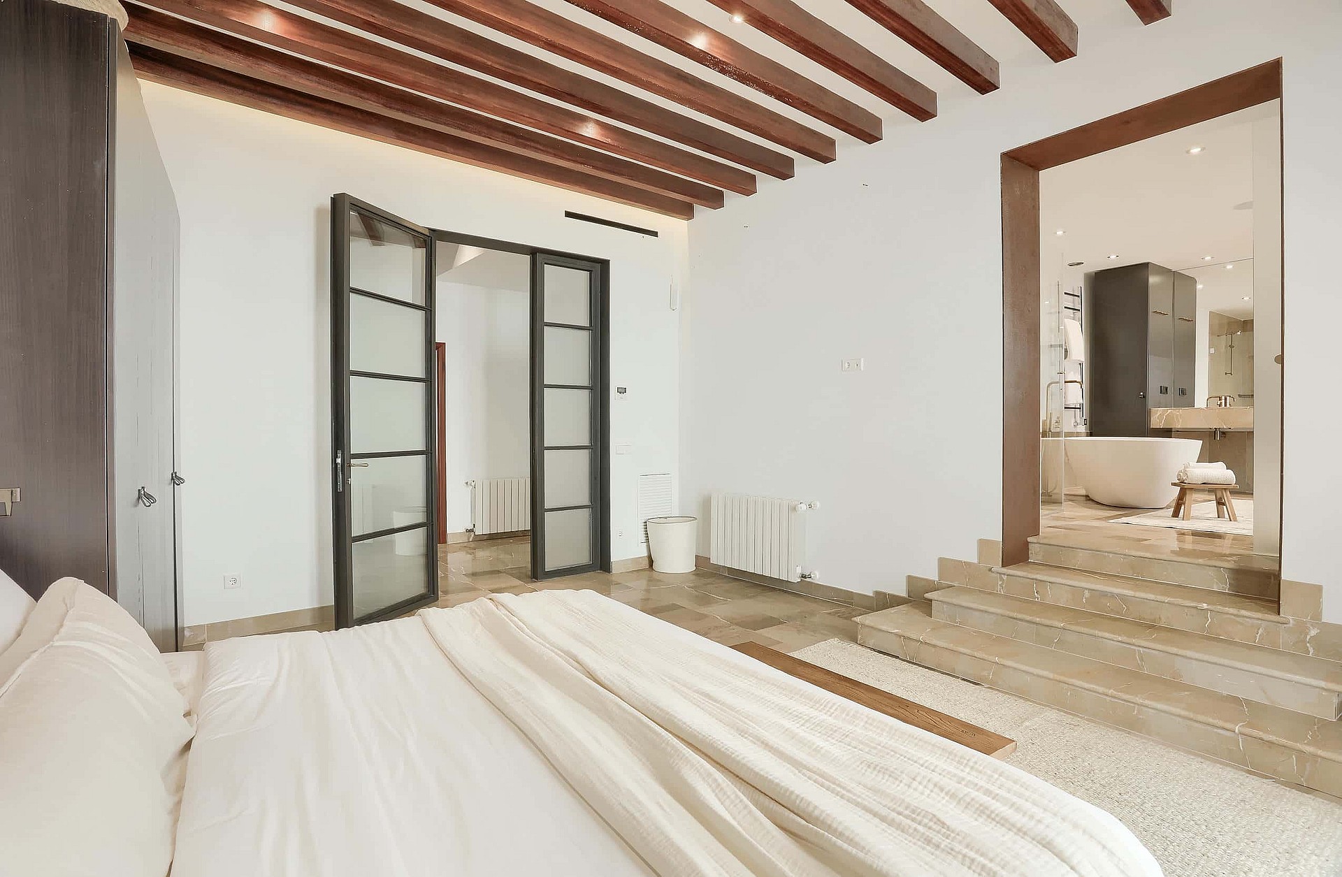 A bright master bedroom with wooden ceiling beams, white bedding, and a raised stone platform leading to an en-suite bathroom.
