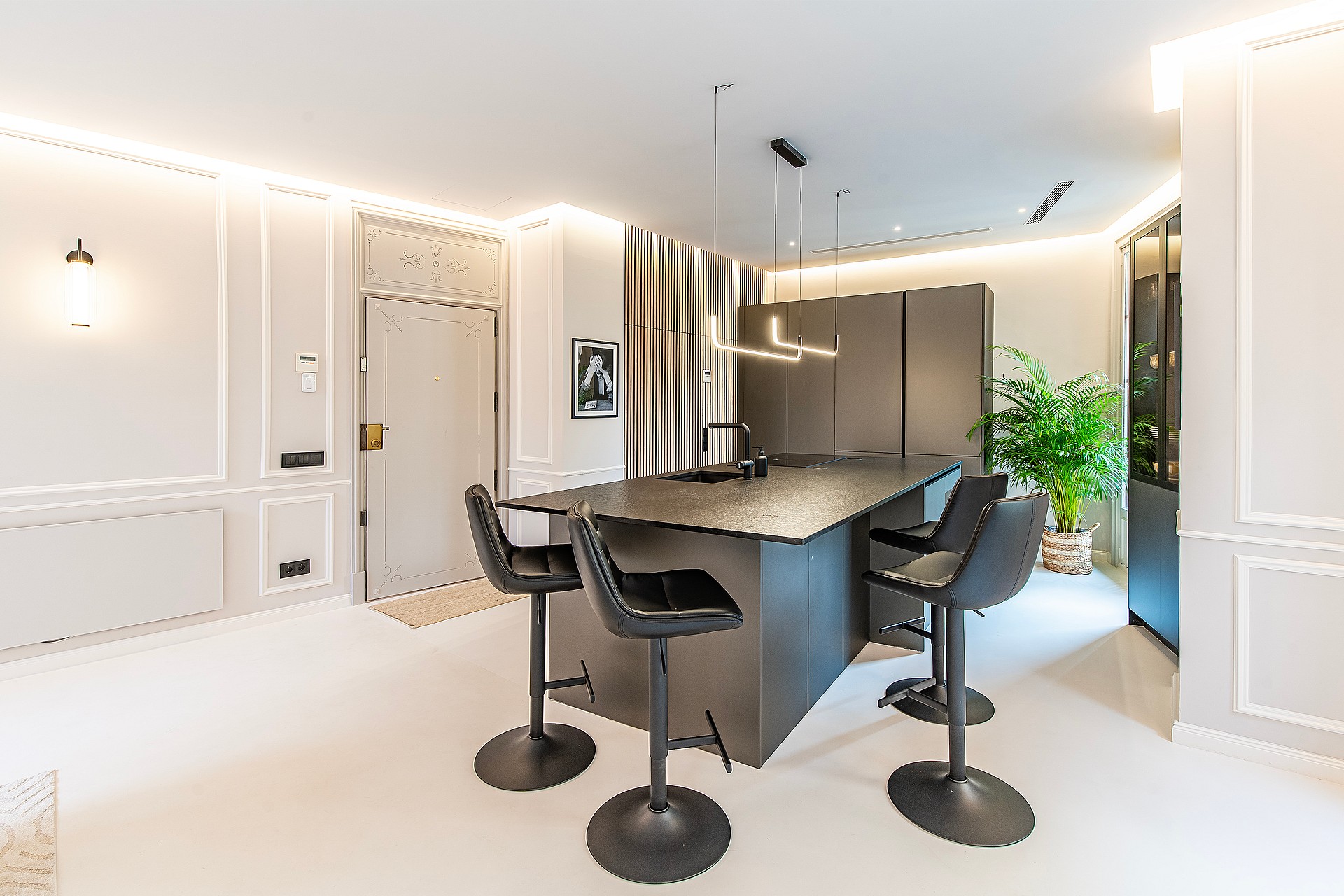 Modern kitchen featuring a dark grey central island, black swivel stools, recessed ceiling lighting, and panelled walls.