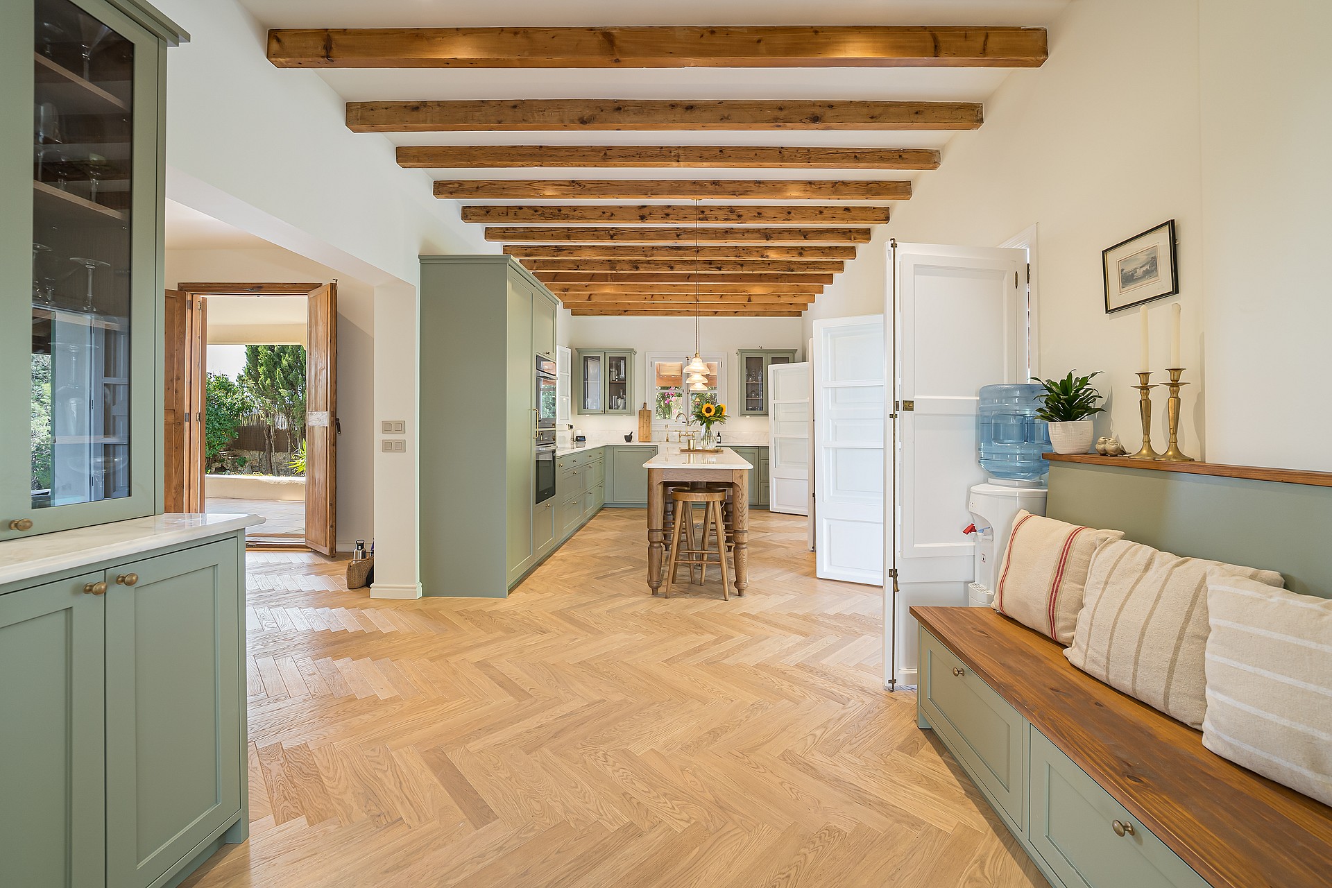 Open-plan kitchen featuring sage green cabinets, light oak herringbone floors, and exposed ceiling beams.