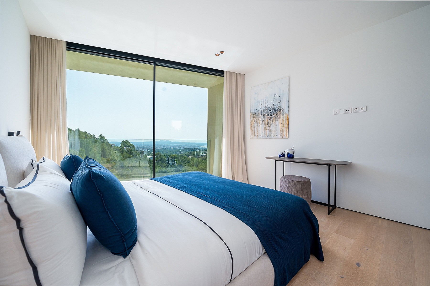Modern bedroom with a large floor-to-ceiling window overlooking a coastal landscape, featuring a white bed with blue pillows.