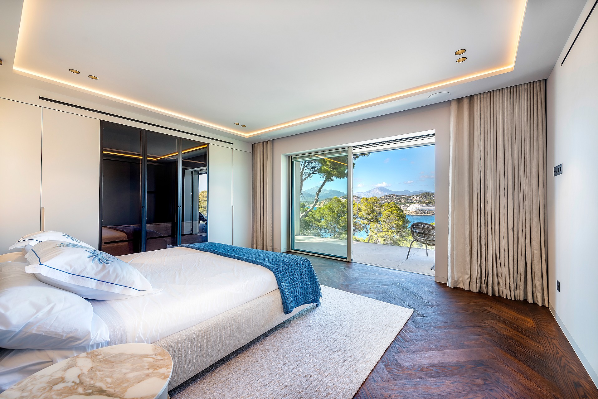 Modern bedroom with dark herringbone wood floors, a large white bed, and floor-to-ceiling glass sliding doors to a terrace.