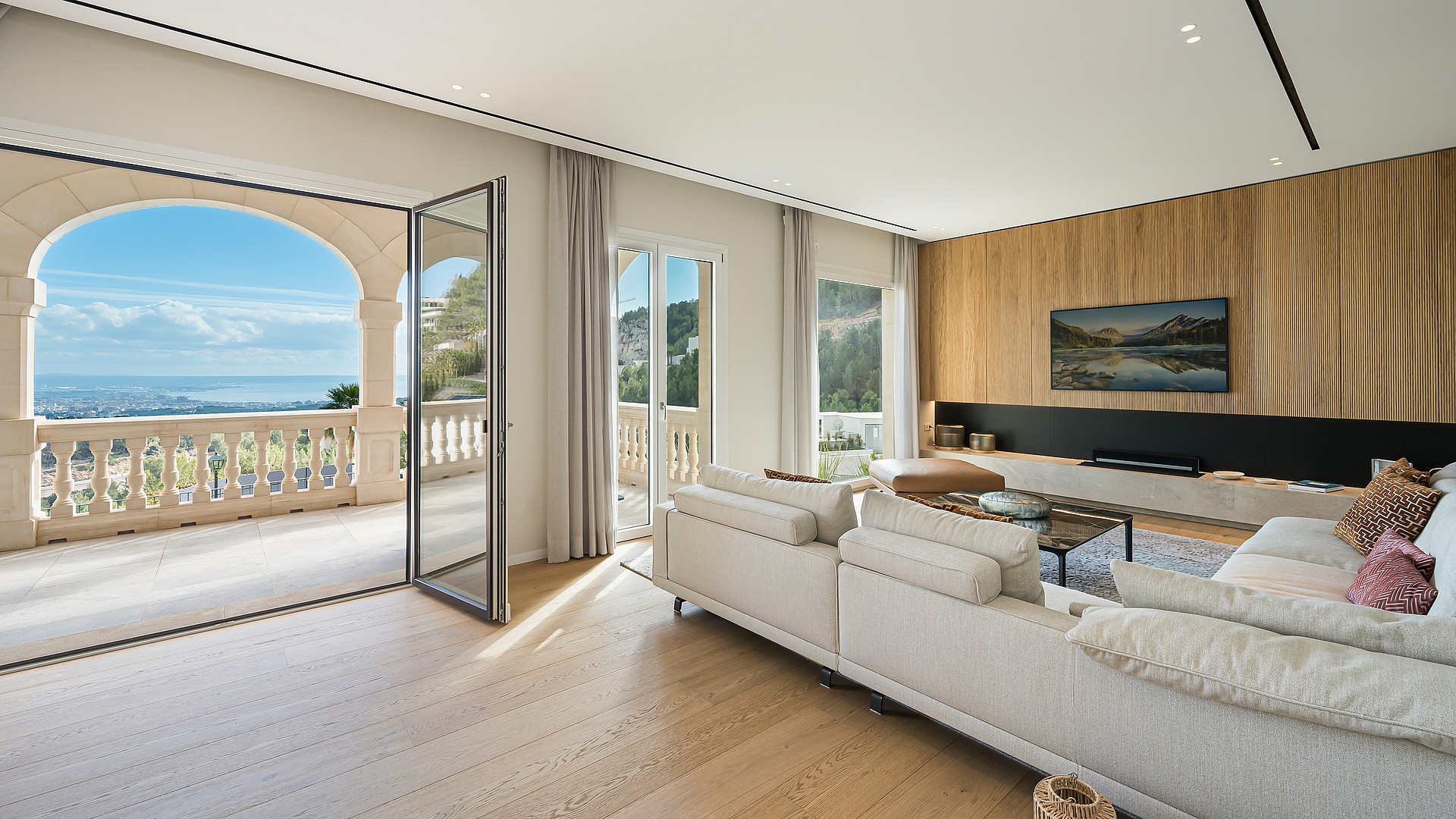 Spacious living room with beige sofa, wooden flooring, and floor-to-ceiling glass doors opening to a stone-railed terrace.