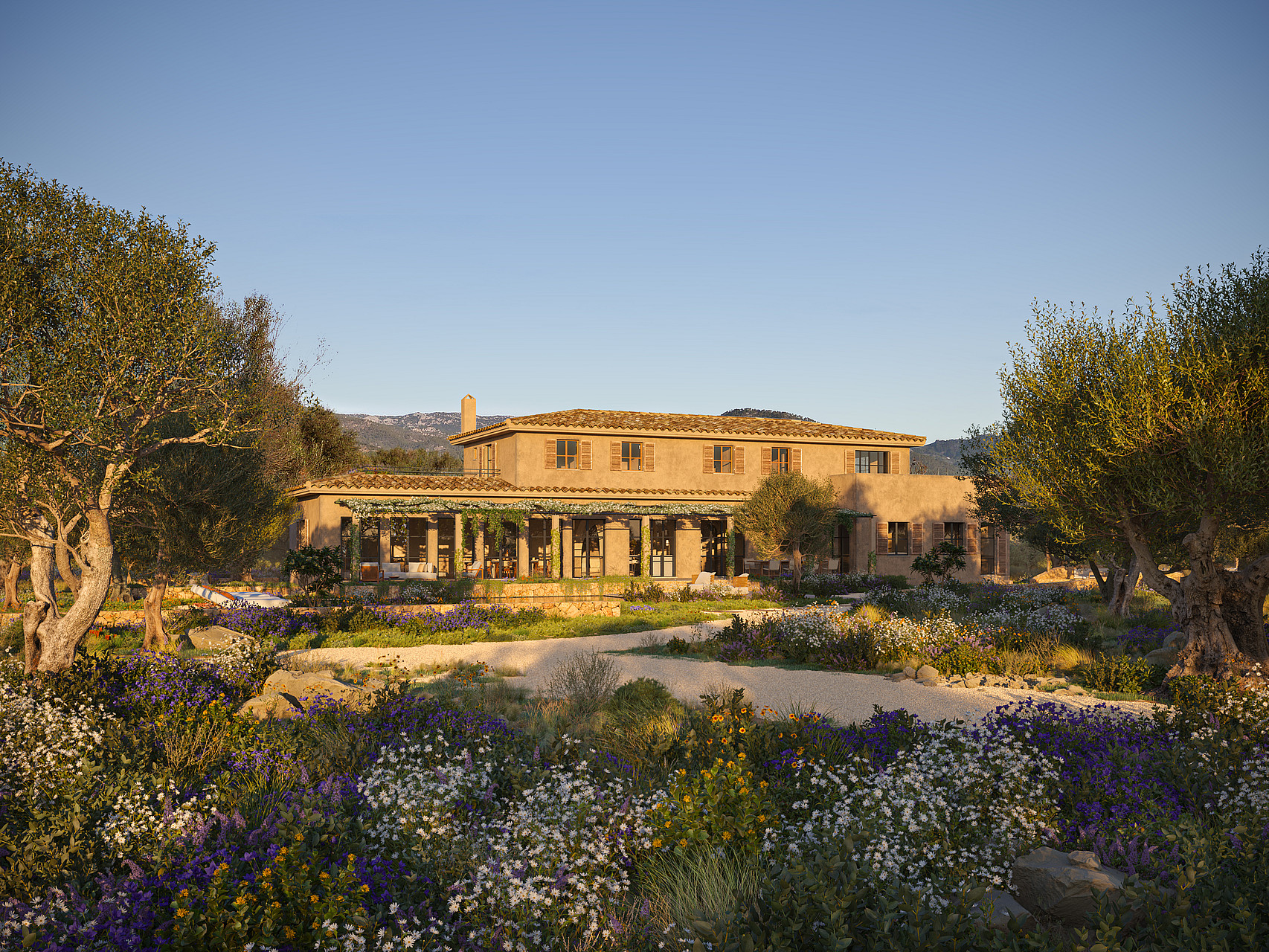 A two-story stone finca set behind a Mediterranean garden with purple wildflowers and olive trees under a clear sky.