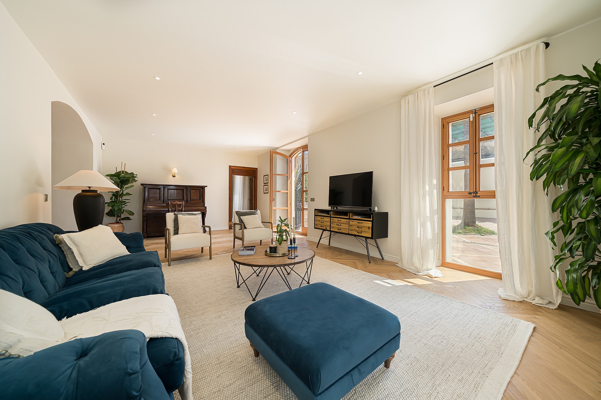 Living area featuring blue velvet tufted sofas, light herringbone wood floors, a dark piano, and arched patio doors.